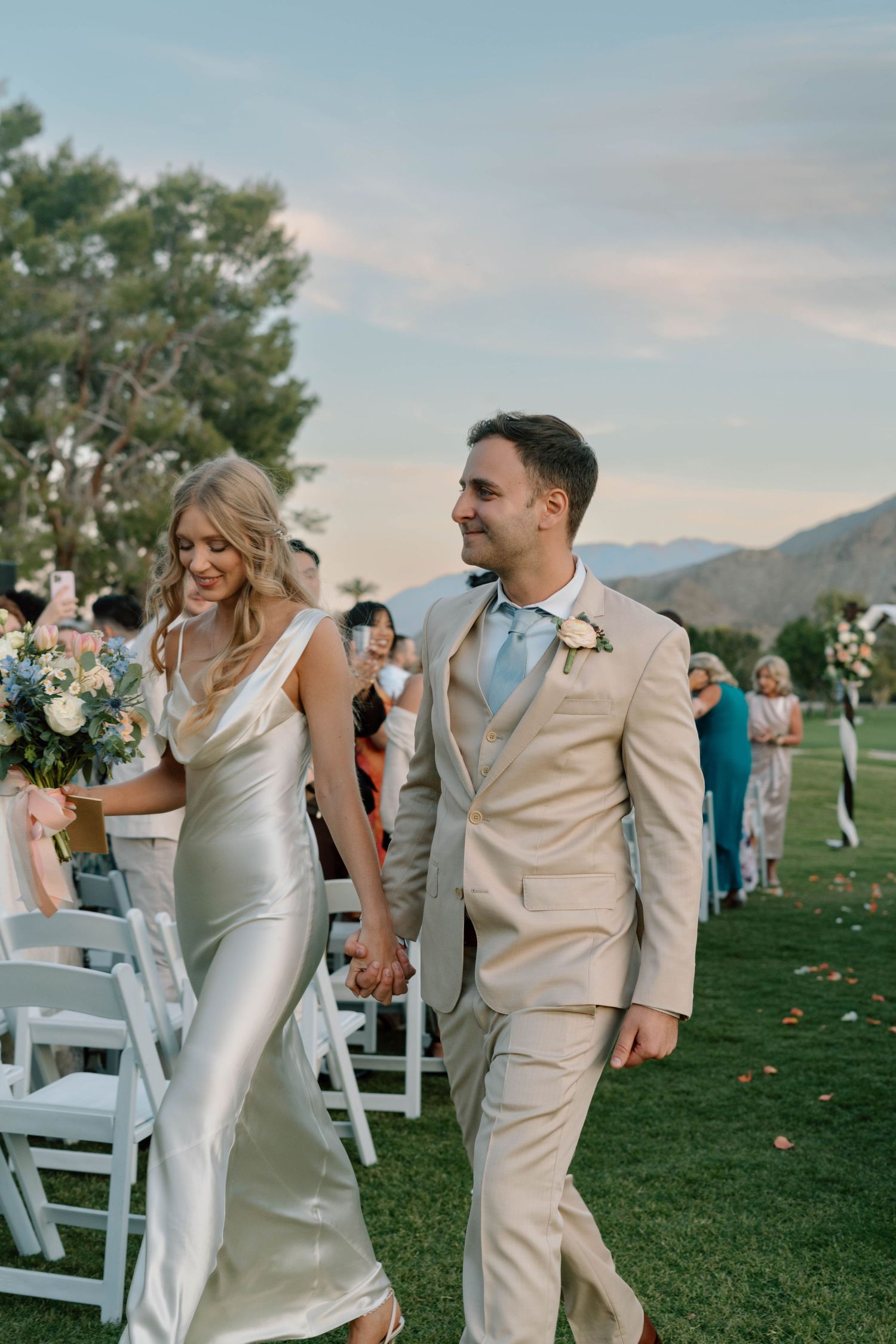 a bride in a white silk dress with a pastel bouque and groom in a tan suit walk down an outdoor aisle during a Palm Springs wedding ceremony