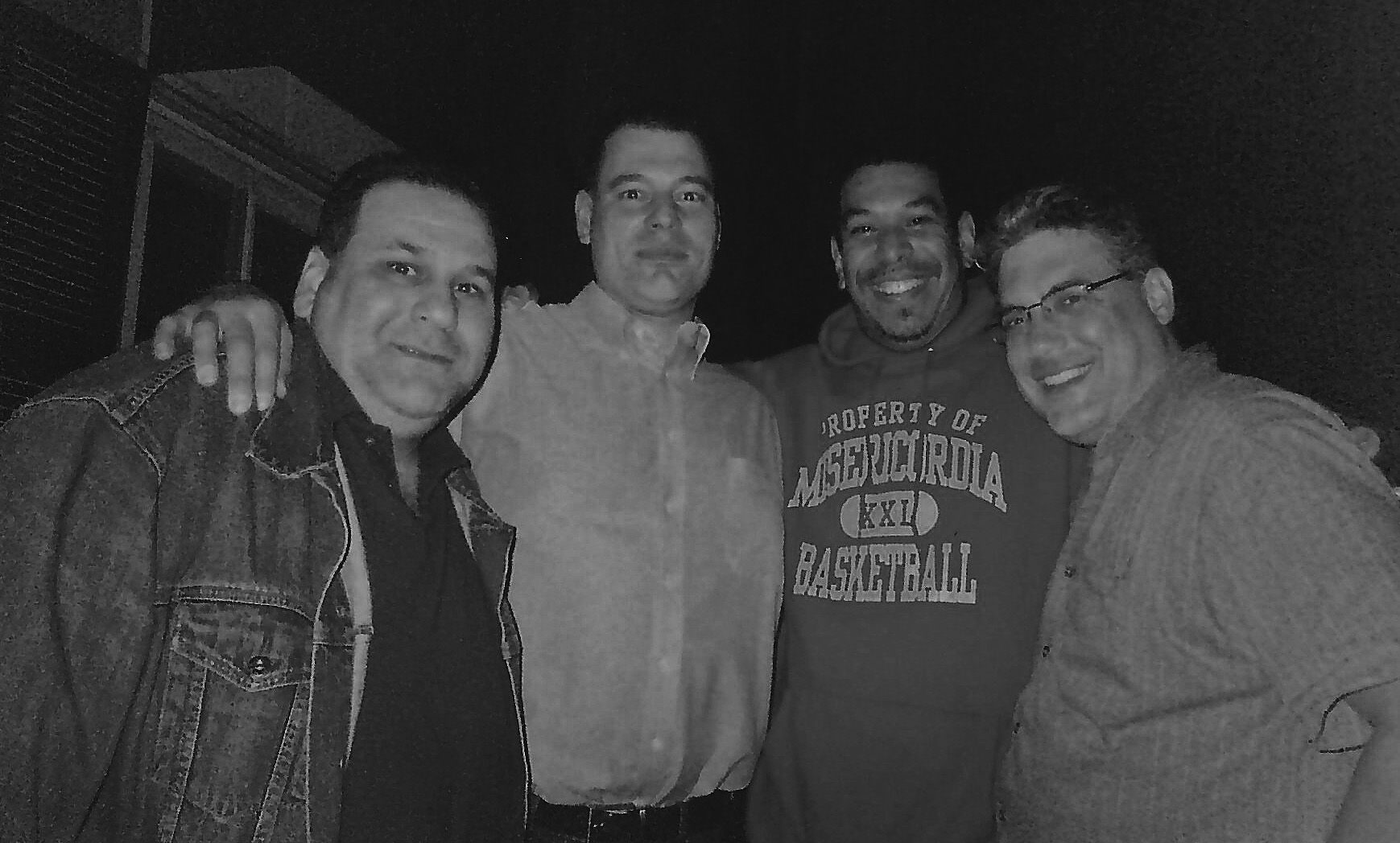 A black and white photograph showing four men standing together in a casual group shot.