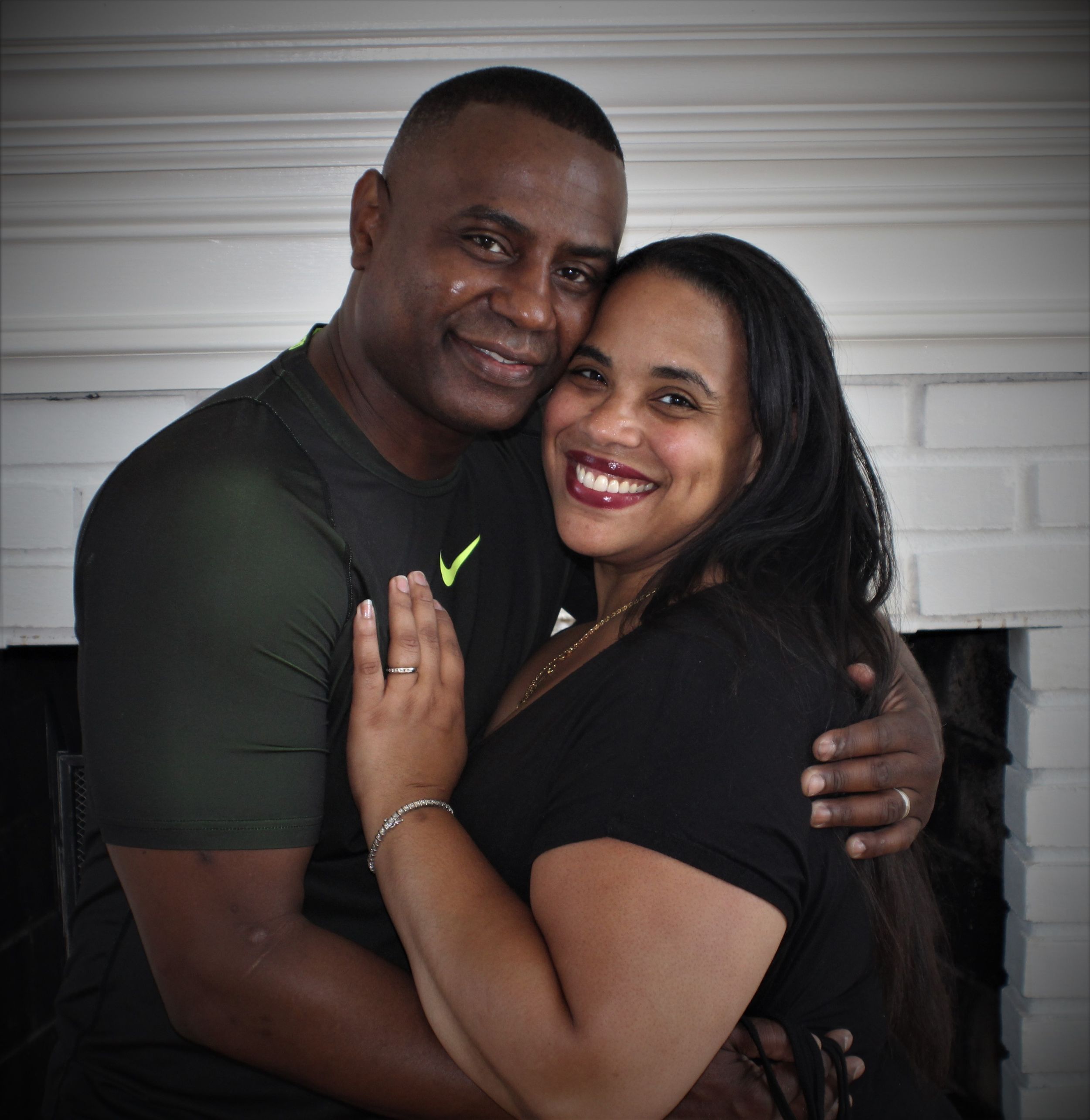 Happy couple in black and green shirts embrace and smile warmly while posing together against a white wall.