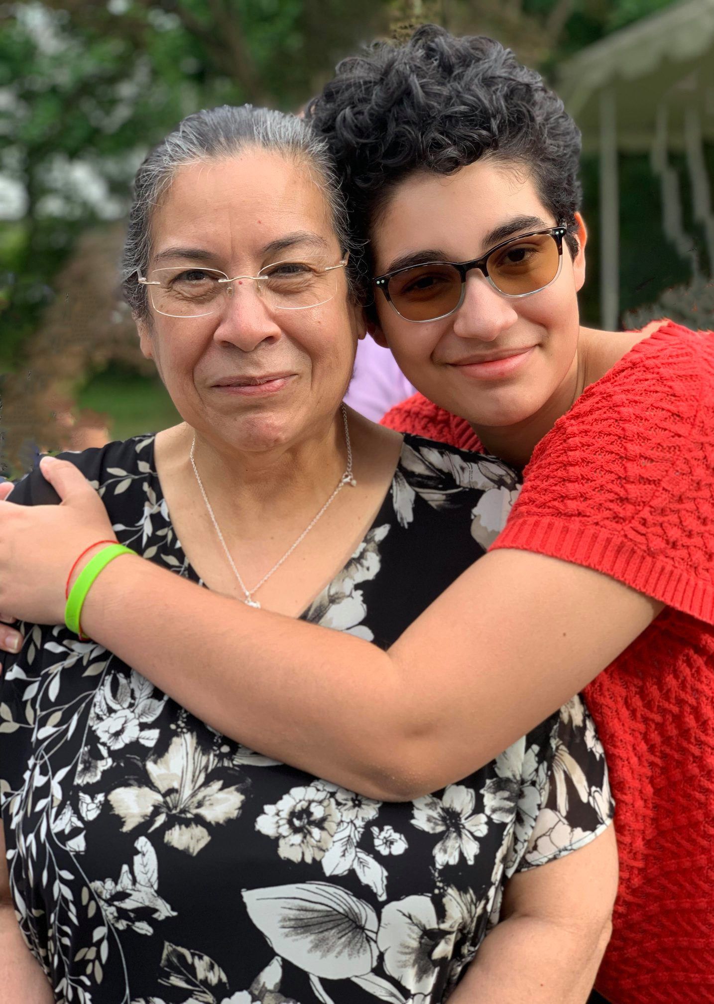 Two individuals wearing glasses embrace and smile outdoors, one in a black floral dress and one in a red sweater.