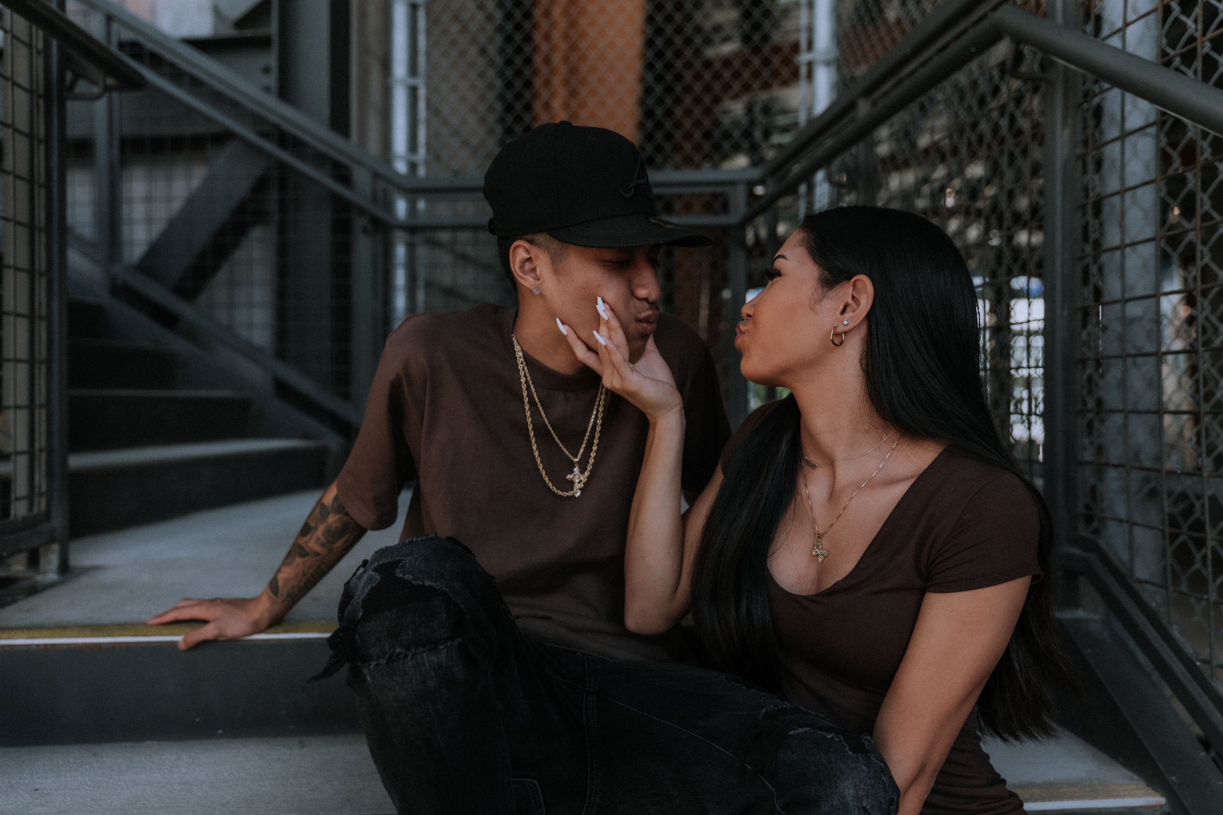 A romantic couple in matching brown outfits share an intimate moment against an industrial background.