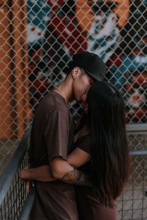 Romantic couple embraces near a chain link fence in matching brown outfits.