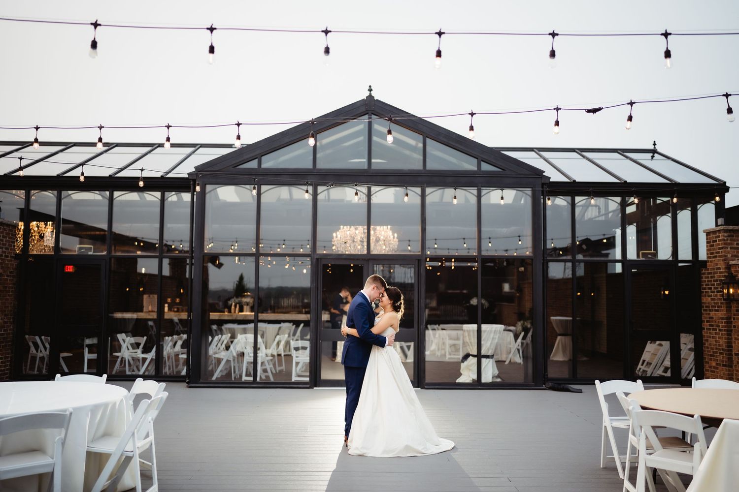 Wedding couple dancing in a modern glass-walled venue with string lights and white chairs.