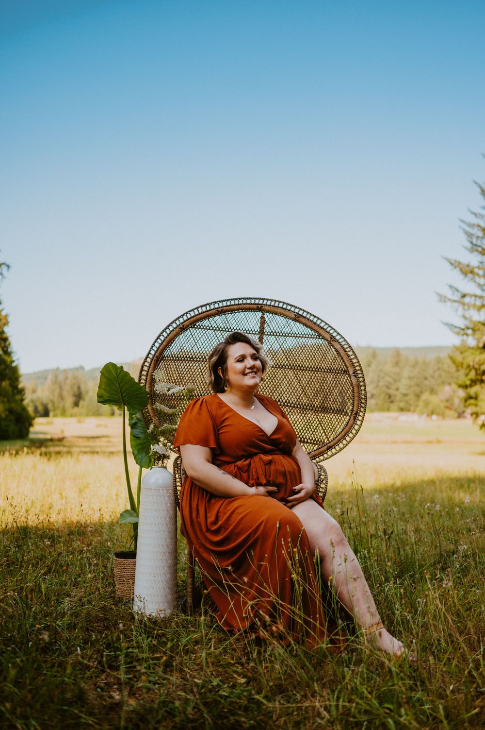 Woman in red dress reclines on vintage peacock chair in sunny meadow setting.