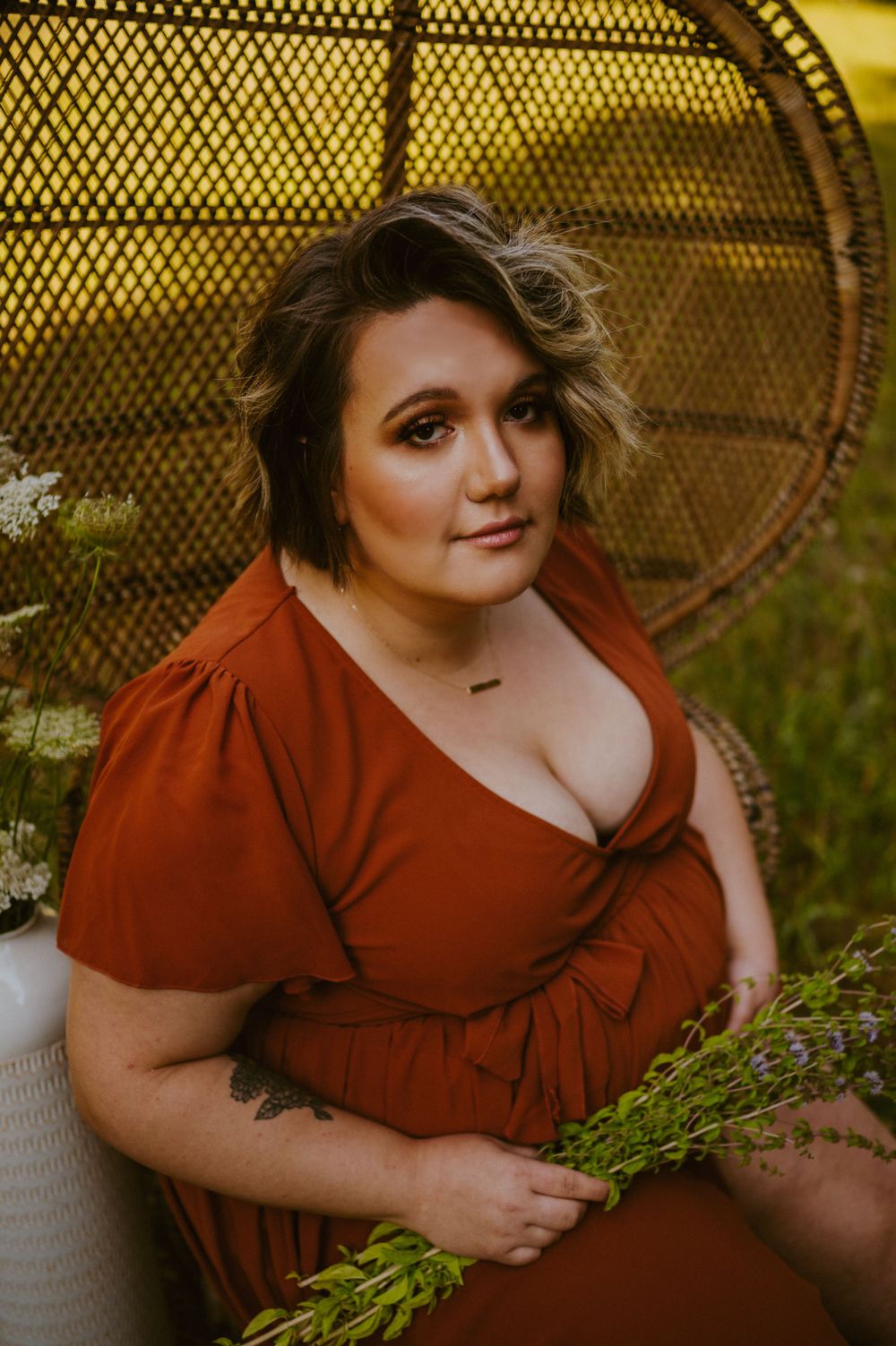 Woman in rust-colored dress poses on peacock chair against yellow mesh background.