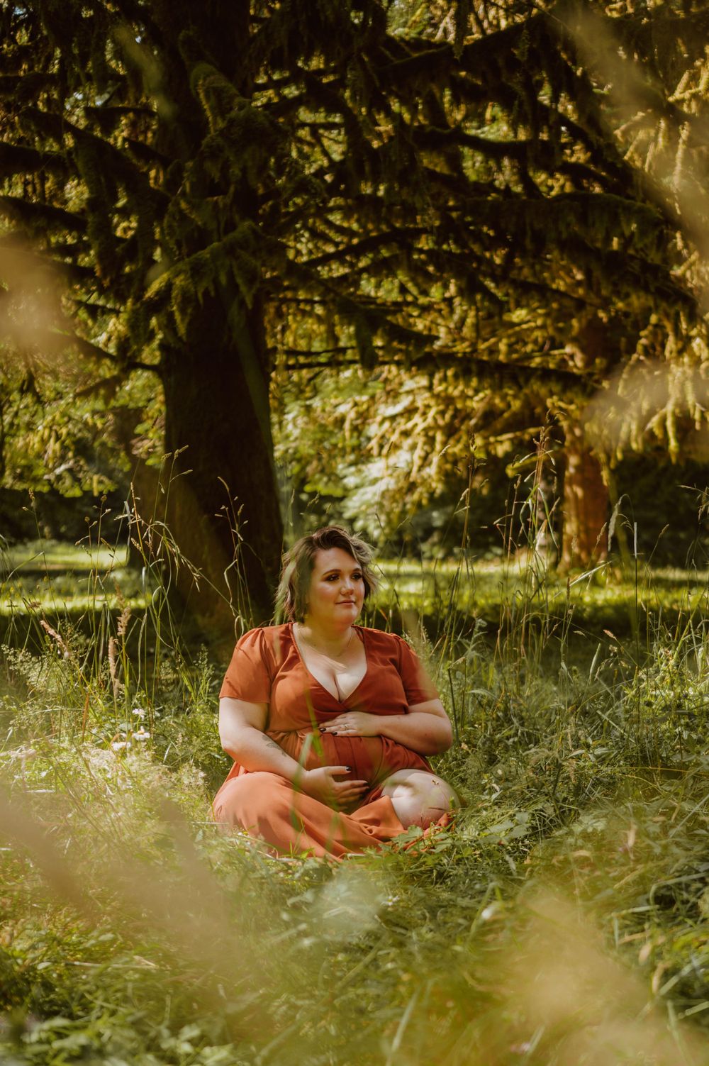 Person in a rust-colored dress sitting peacefully in a sunlit grassy area under trees.