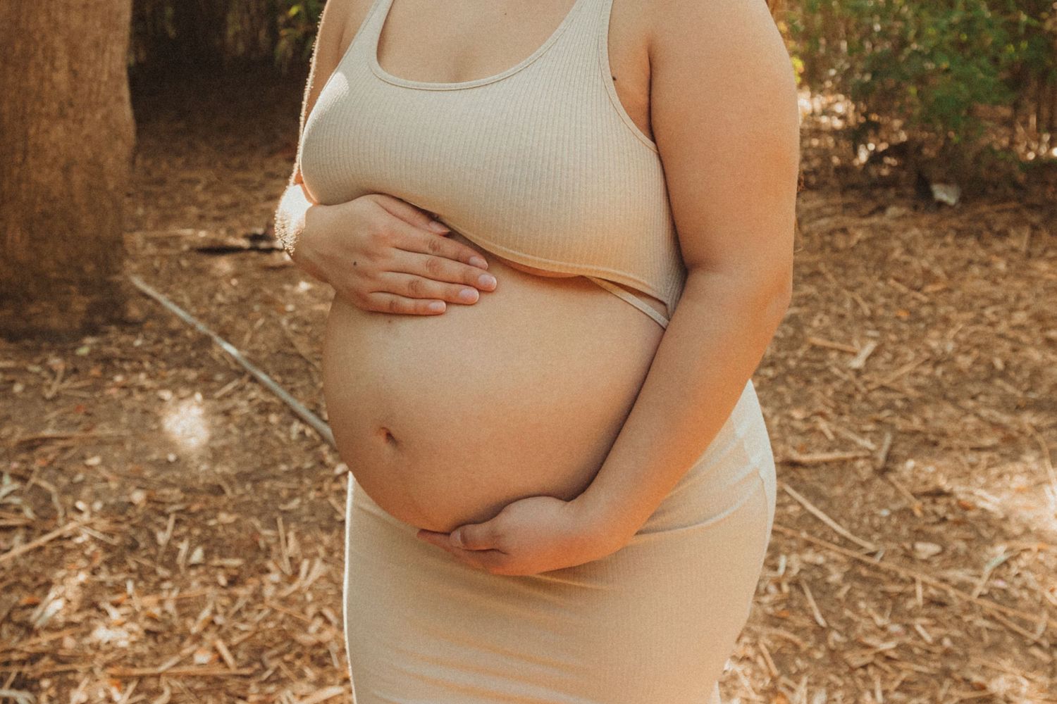 Maternity photo showing pregnant belly in beige tank top against natural outdoor backdrop.