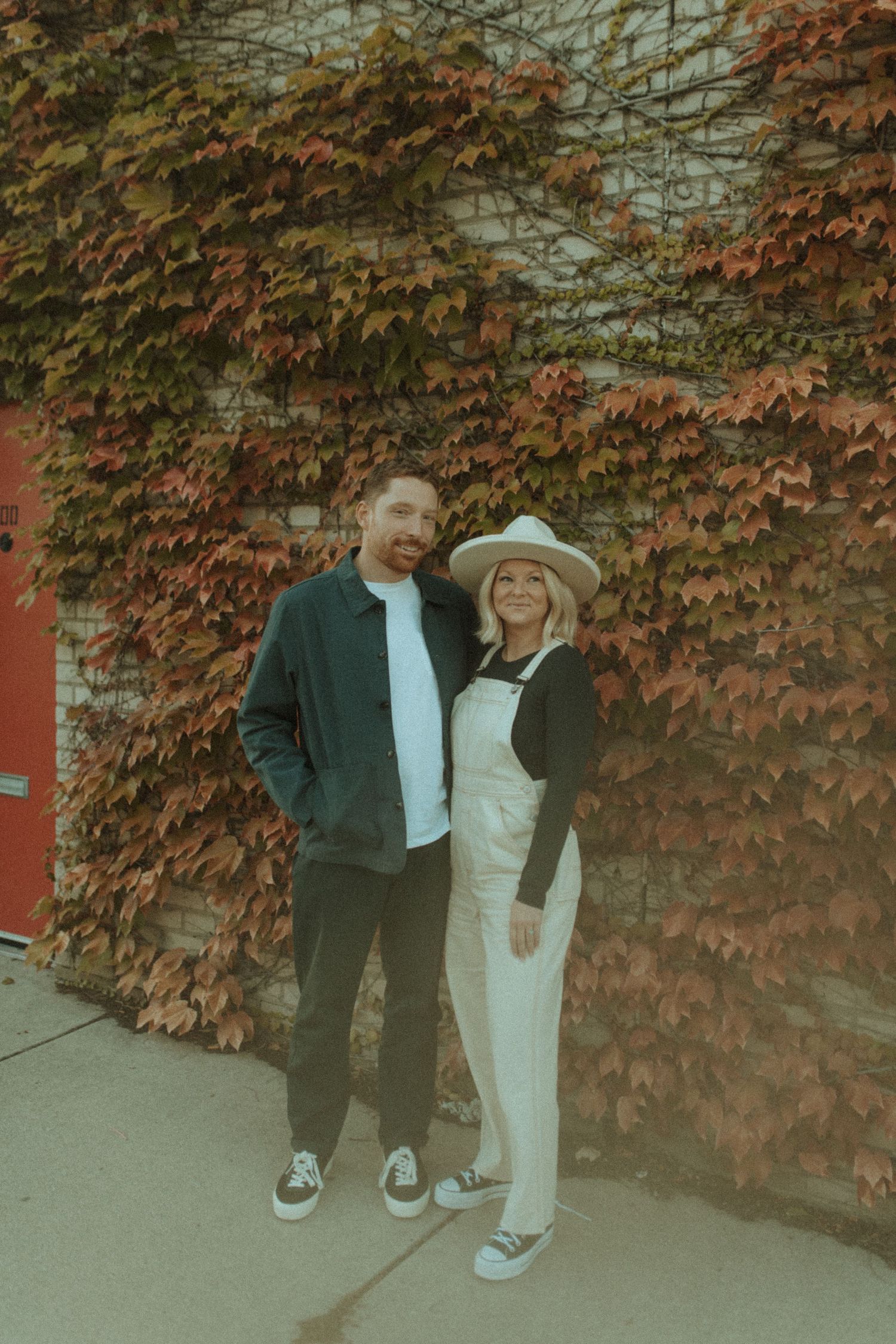 A couple smiles in front of a brick wall covered in autumn ivy wearing complementary casual outfits.