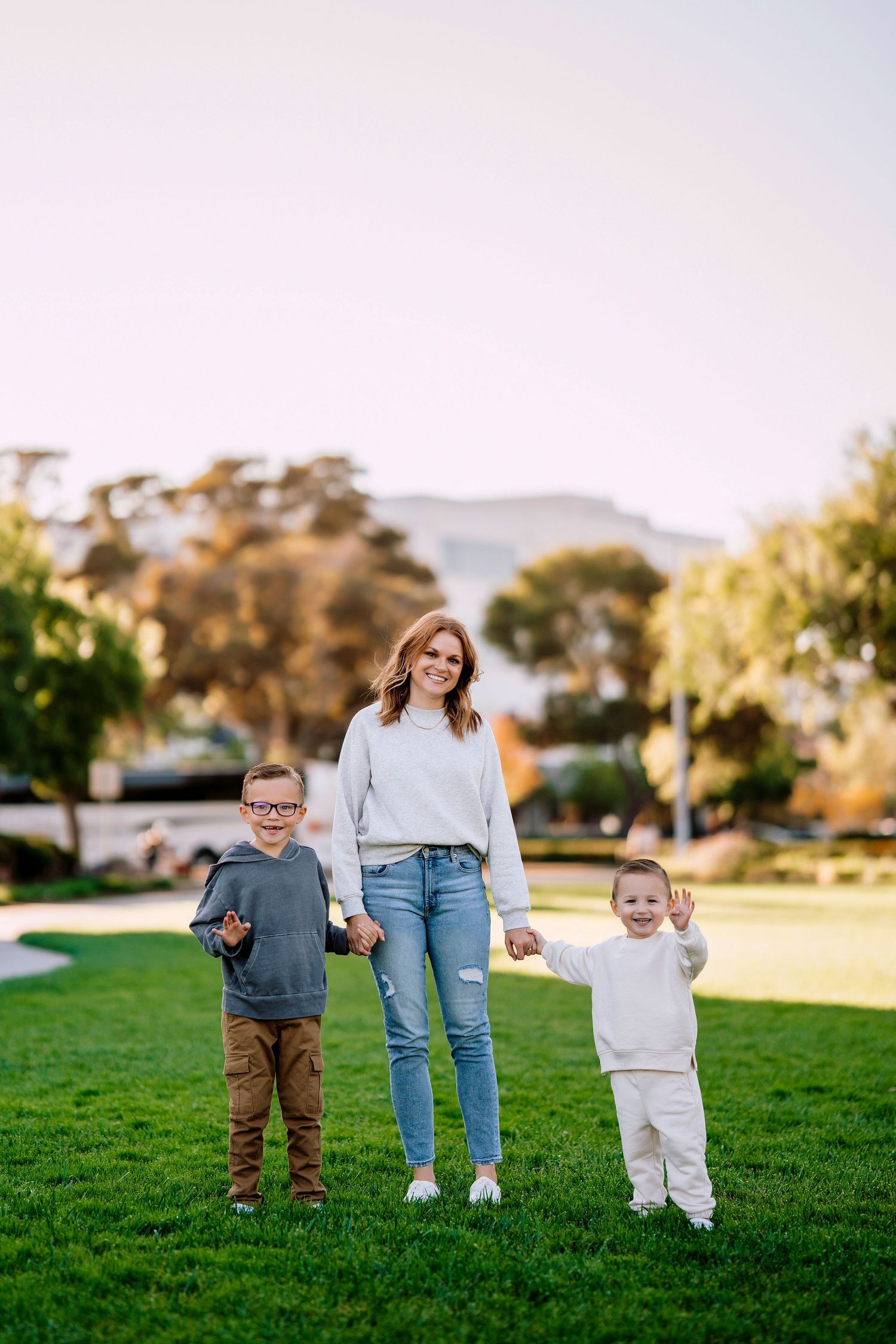 A family holds hands and stands together on a grassy lawn during golden hour at a park.