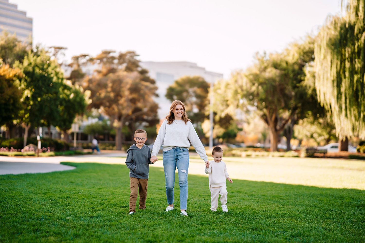 Three people walk hand in hand through sunny park with trees in background.