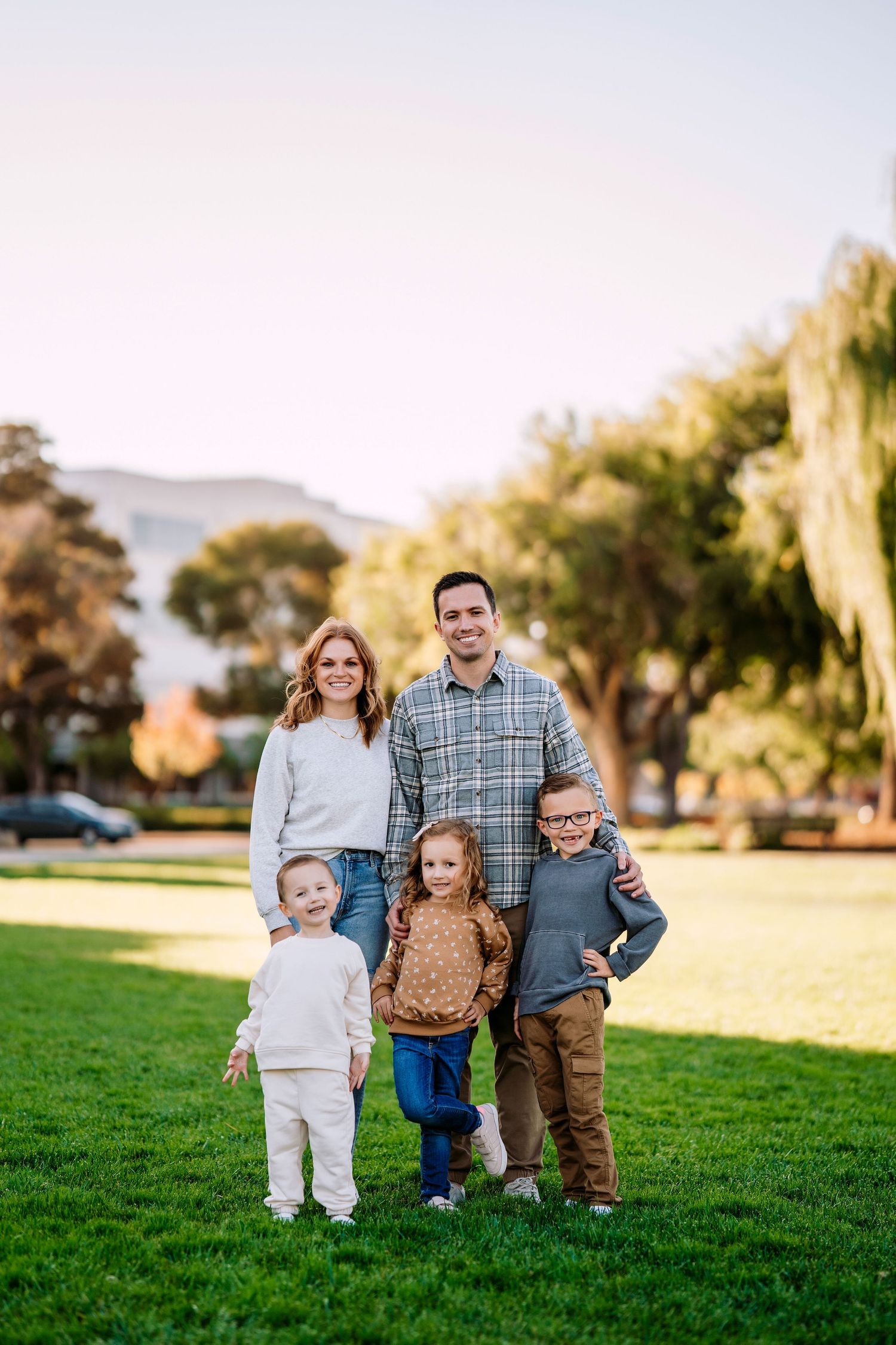 A family of five poses together on a grassy lawn with weeping willow trees and mountains in the background.