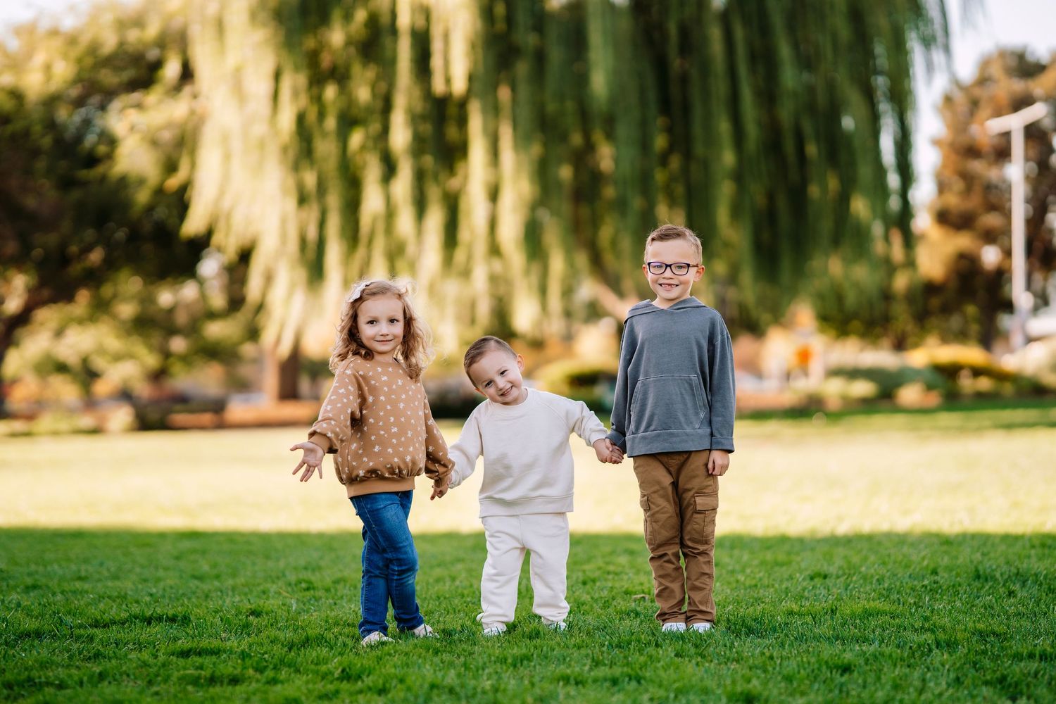 Three children stand together holding hands and playing on a green lawn beneath a weeping willow tree.