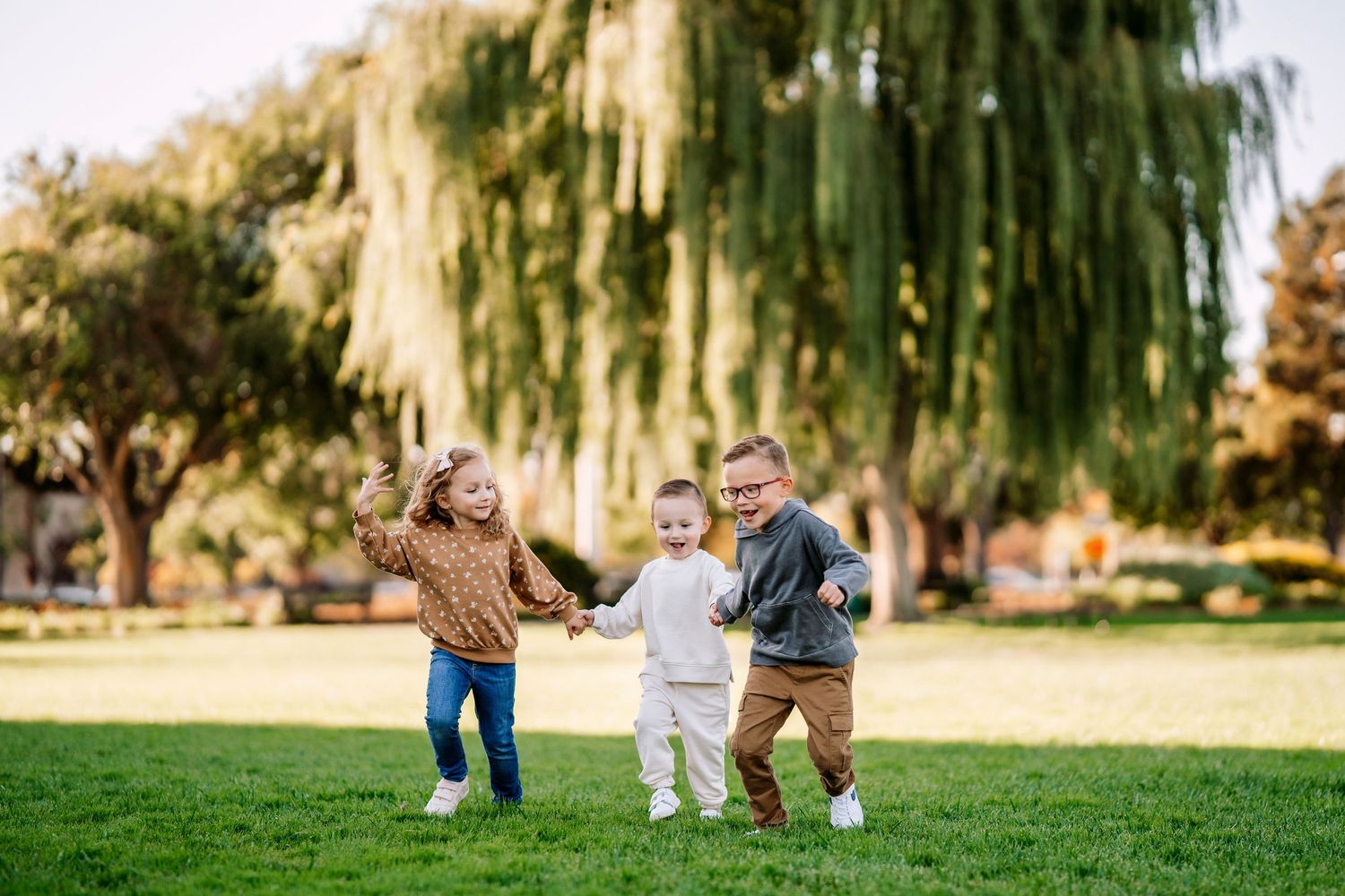 Three children run and play together on grass under a large weeping willow tree.