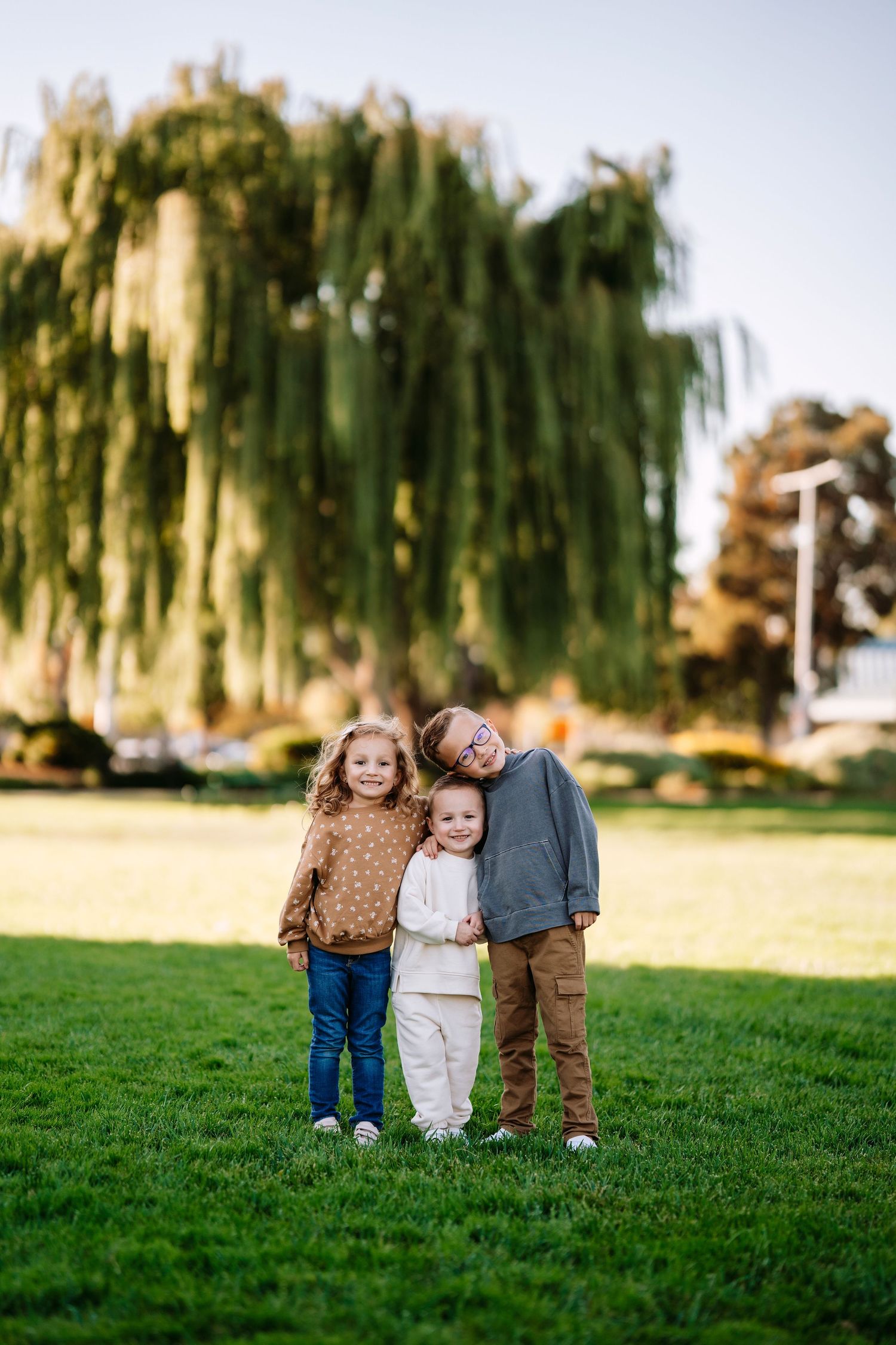 Three siblings pose together on grass with a scenic willow tree backdrop during golden hour.