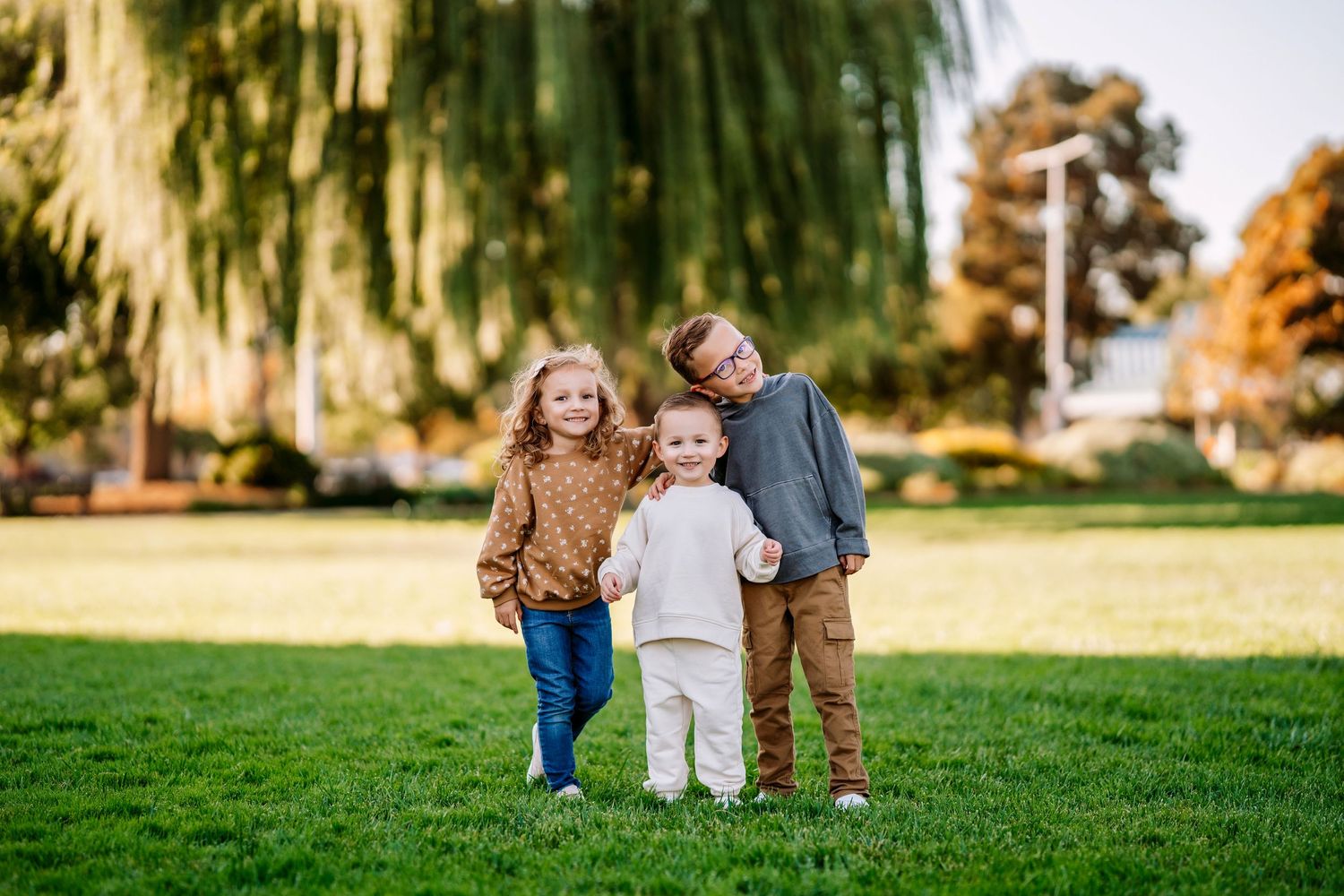 Three young children stand close together on grass with a willow tree in the background.
