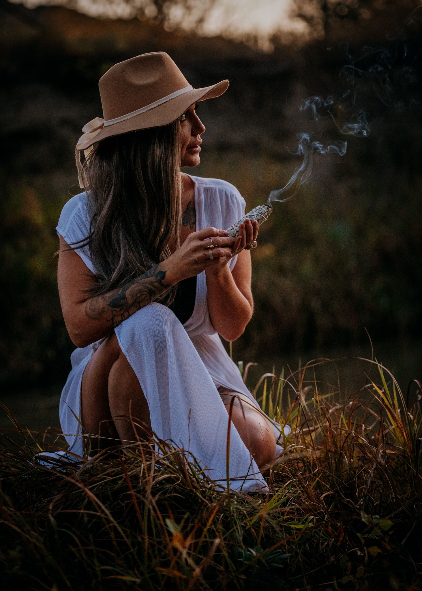 Woman in white dress and brown hat sits in field at sunset with smoke wisping from hand.