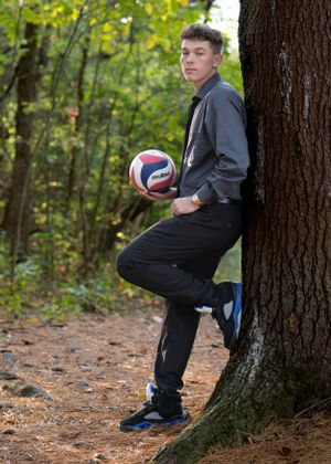 Athletic person in dark clothes poses with volleyball against tree in forest setting.
