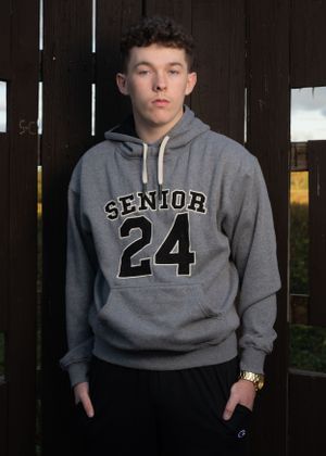 Person wearing grey Senior 24 hoodie posing against dark wooden fence.