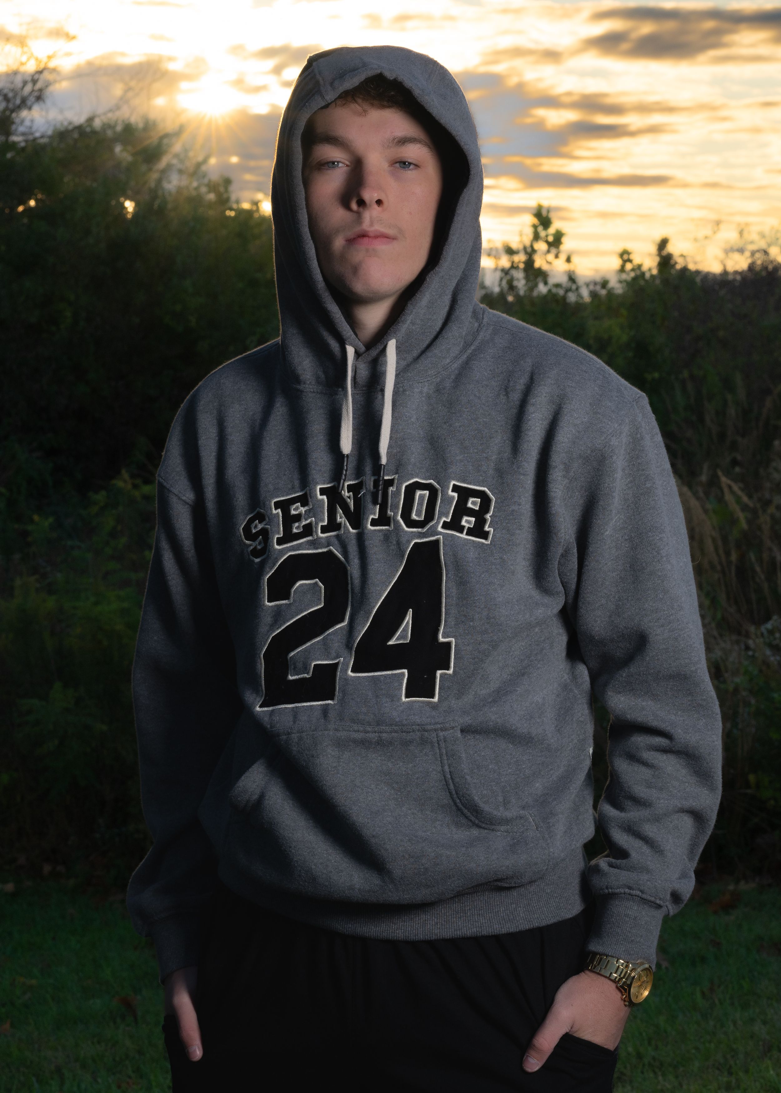 Person in grey Senior 24 hoodie poses at sunset outdoors.