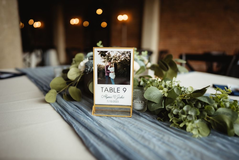 Table number display framed in gold with eucalyptus greenery and blue table runner at wedding reception.