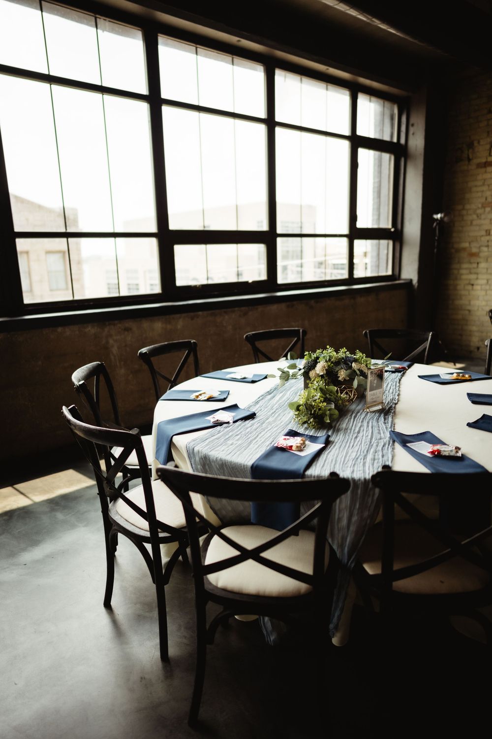 Modern reception table setting with navy napkins near large industrial windows.