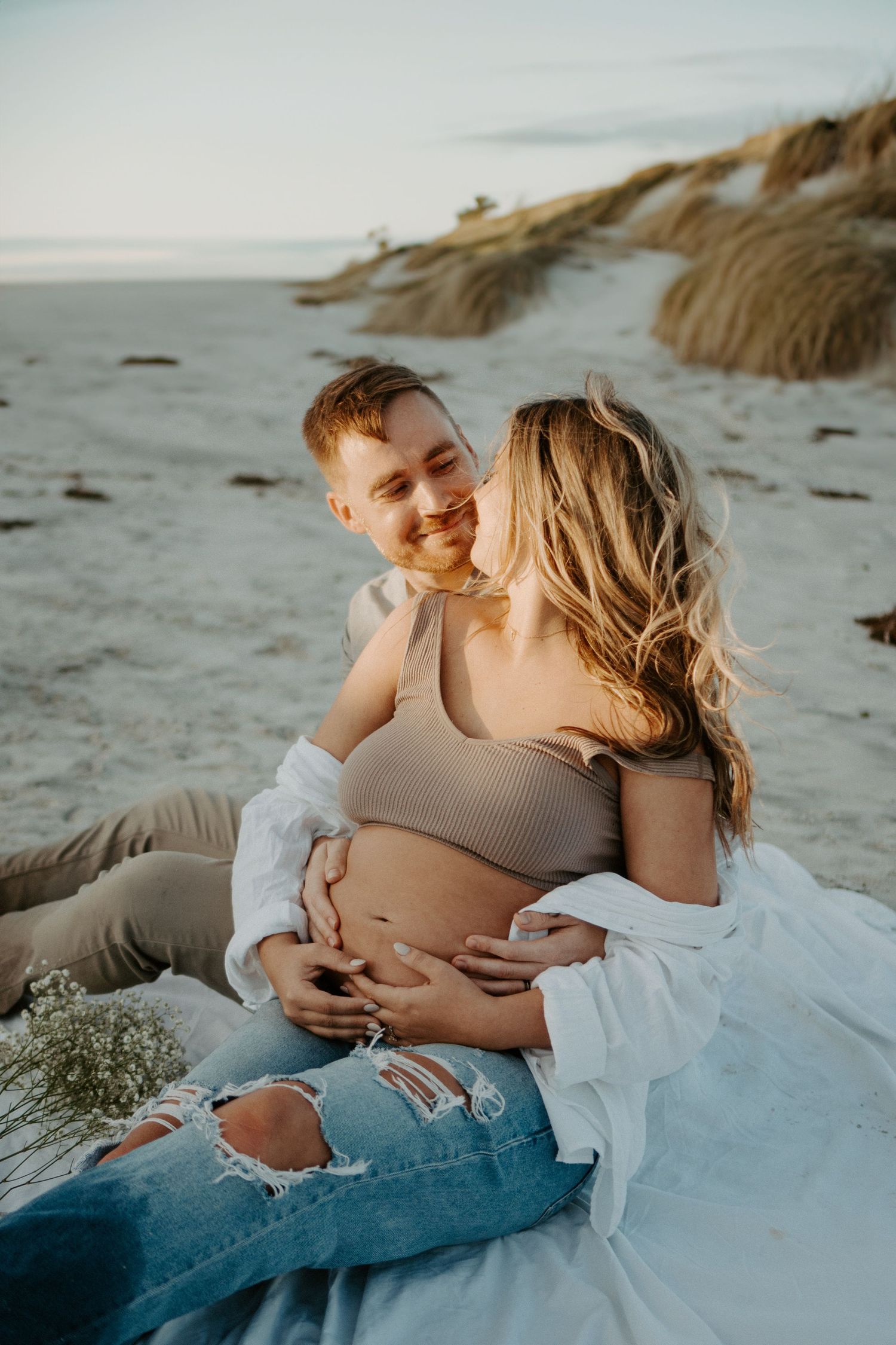 Maternity Couple posed sitting in the sand with the ocean and sand dunes in the background
