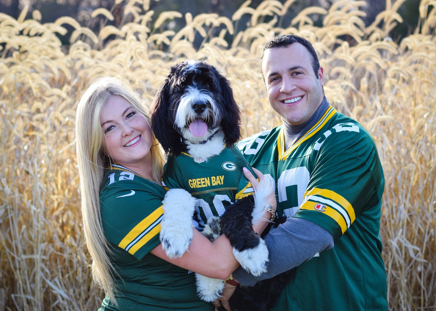 Couple in Green Bay Packers jerseys holds their black and white dog in a cornfield.