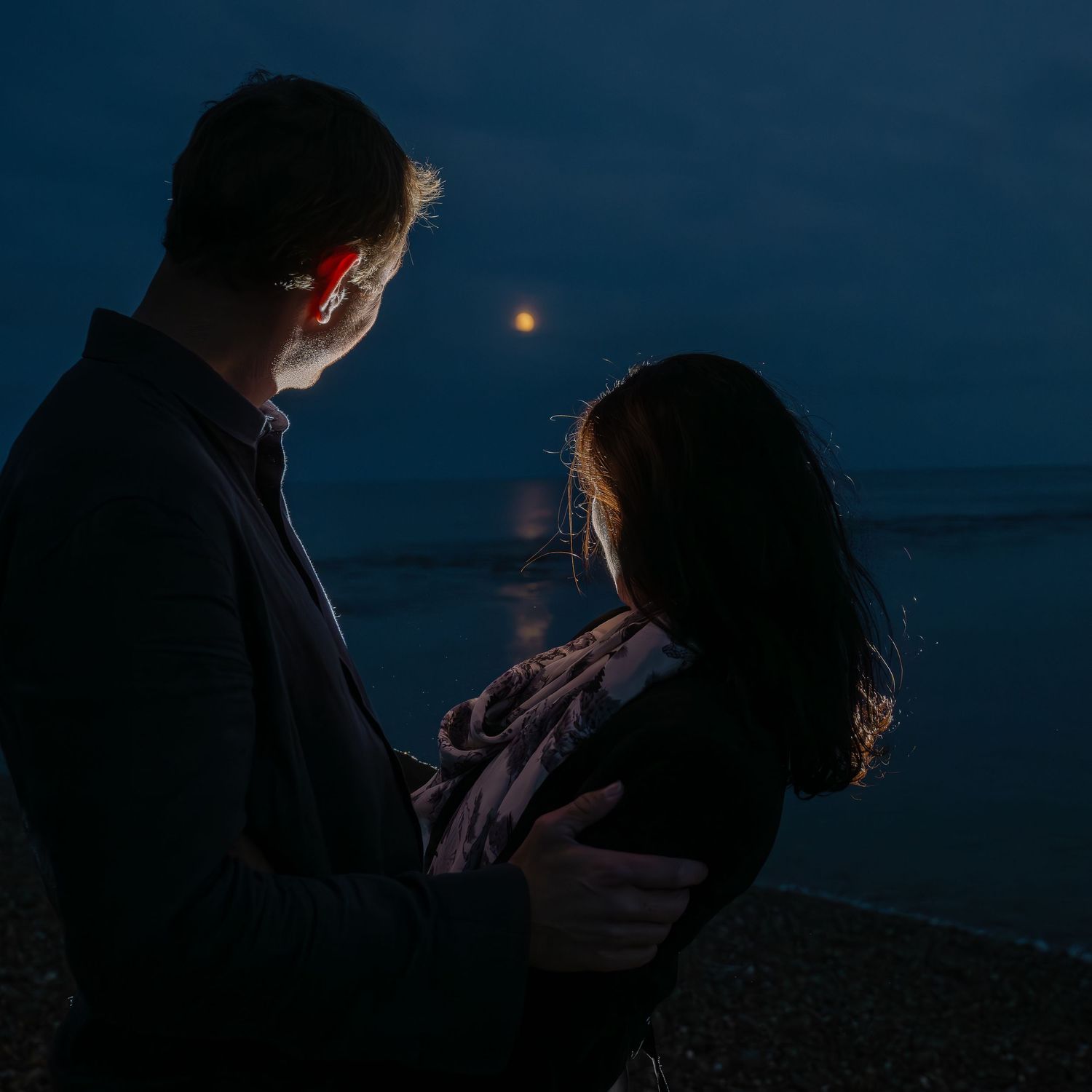Silhouetted couple shares intimate moment on beach at night with moon reflection.