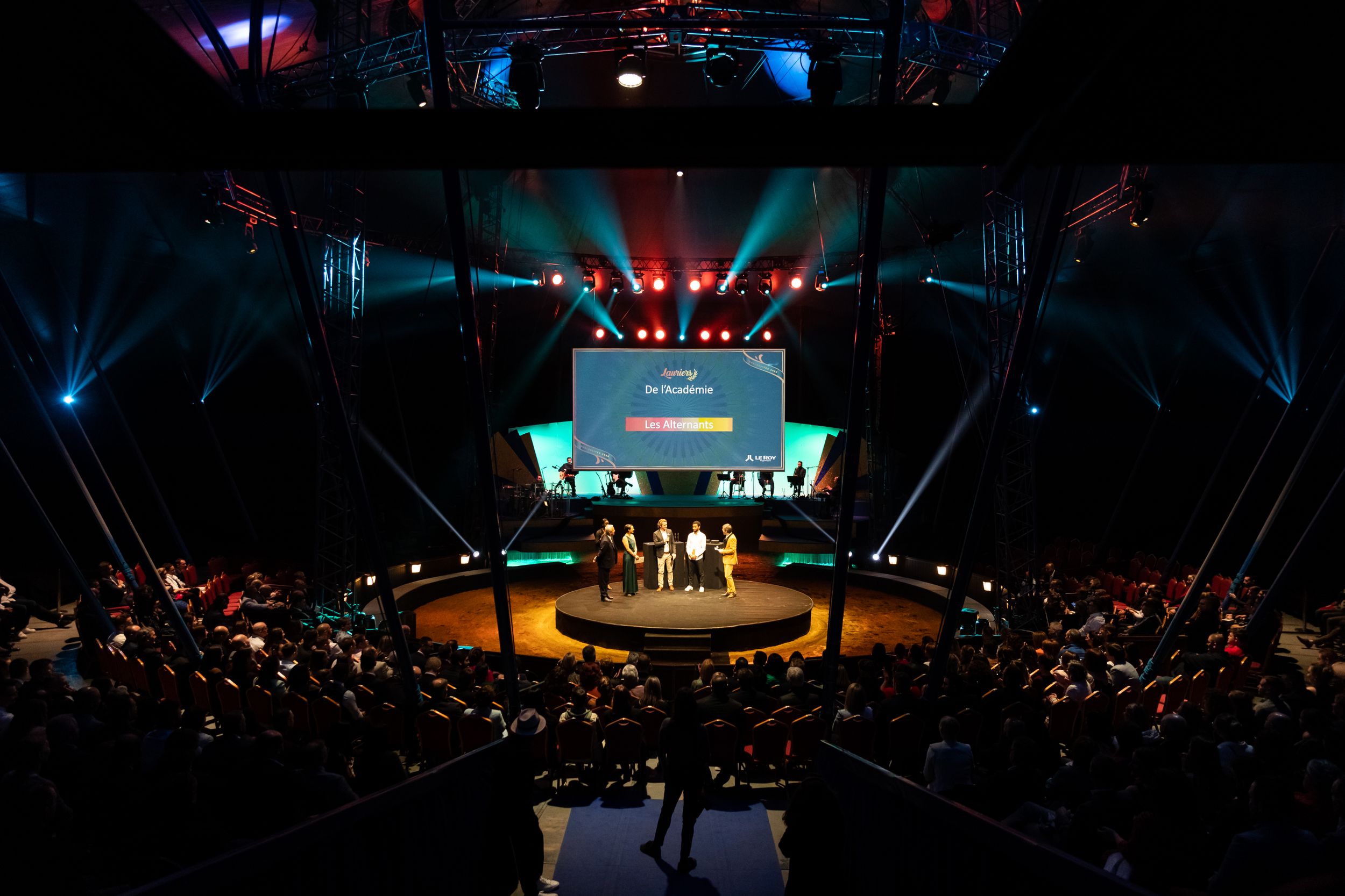Large indoor event space with circular stage and audience seating illuminated by blue and red spotlights.