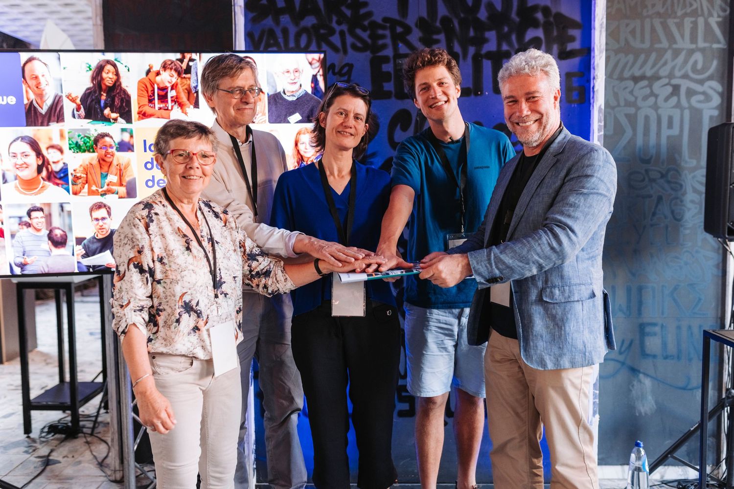 Group joins hands in unity pose at business presentation with photo wall display behind them.
