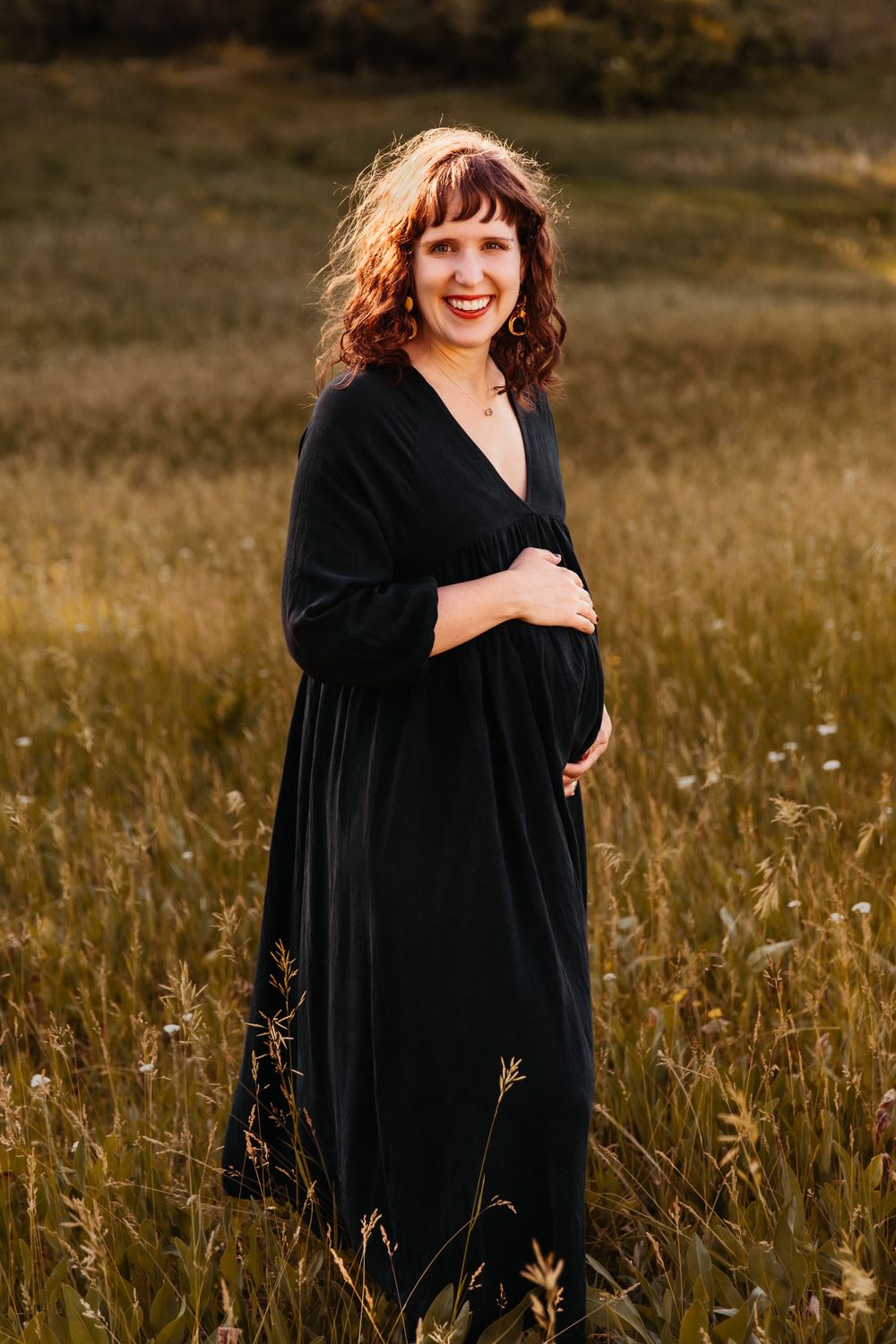 Pregnant woman with curly brown hair and long hunter green dress stands in a field of tallgrass.