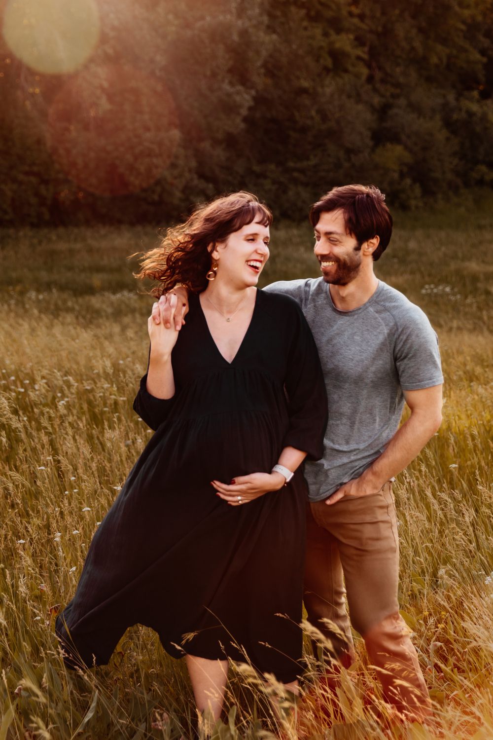Pregnant woman stands next to her husband in a field during golden hour. They're looking at each other and laughing.