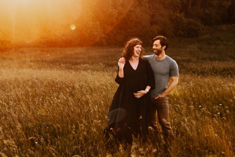 Pregnant woman and her husband laugh together in a field of tallgrass. The orange sun glows behind them.