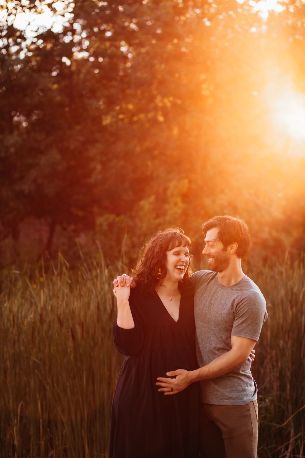 Pregnant woman shares in laughter with her husband. His had is on her baby bump and the golden sun glows behind them.