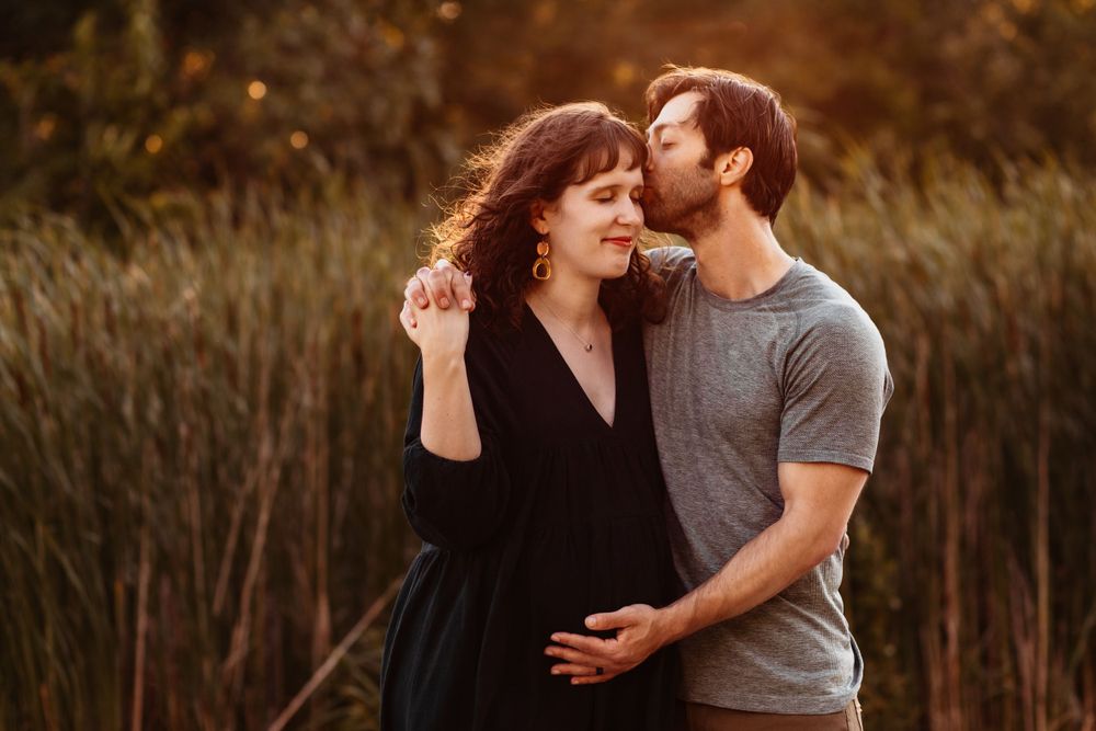 A man kisses the temple of his pregnant wife. One hand clasps onto her shoulder while the other touches her baby bump.