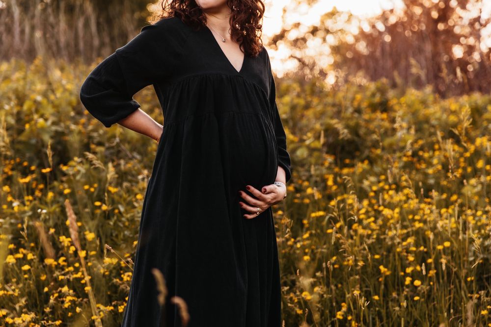 Pregnant woman in a flowing dark green dress stands amongst a field of yellow flowers.
