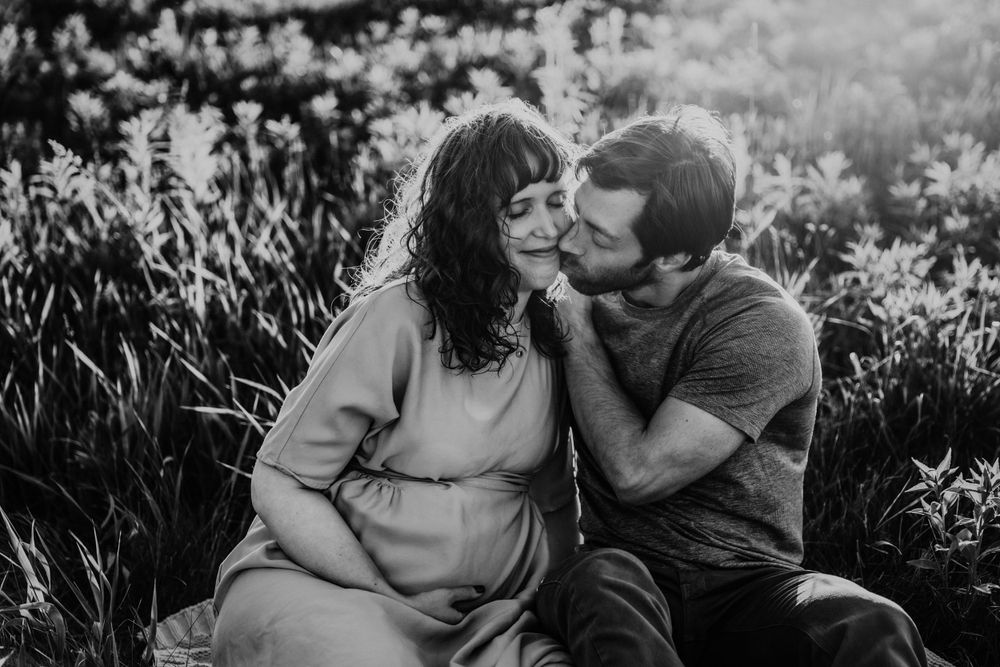 Black-and-white image of a husband kissing his pregnant wife's cheek during golden hour.