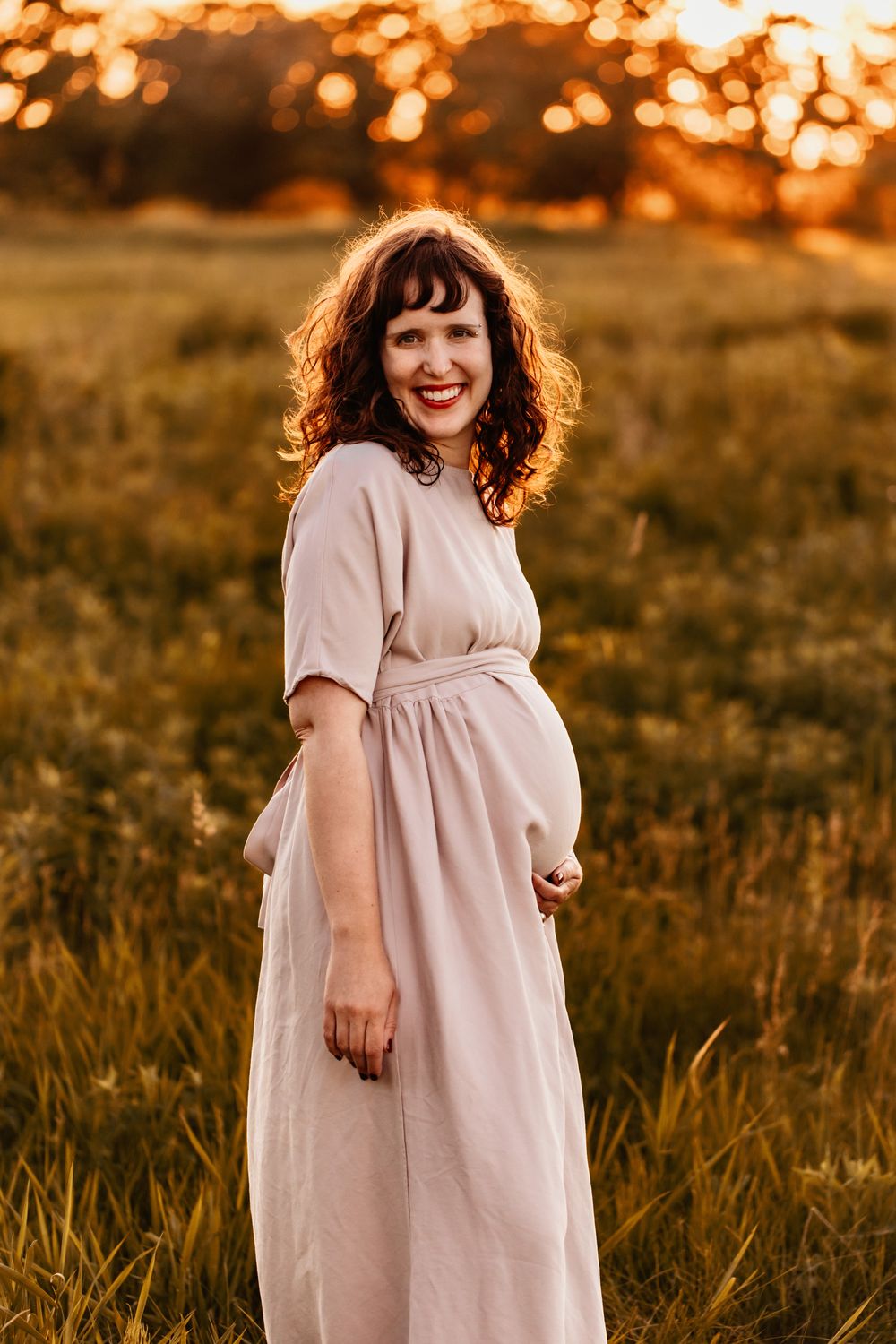 Pregnant woman smiles at the camera in a field of green during golden hour. One hand rests on her baby bump.