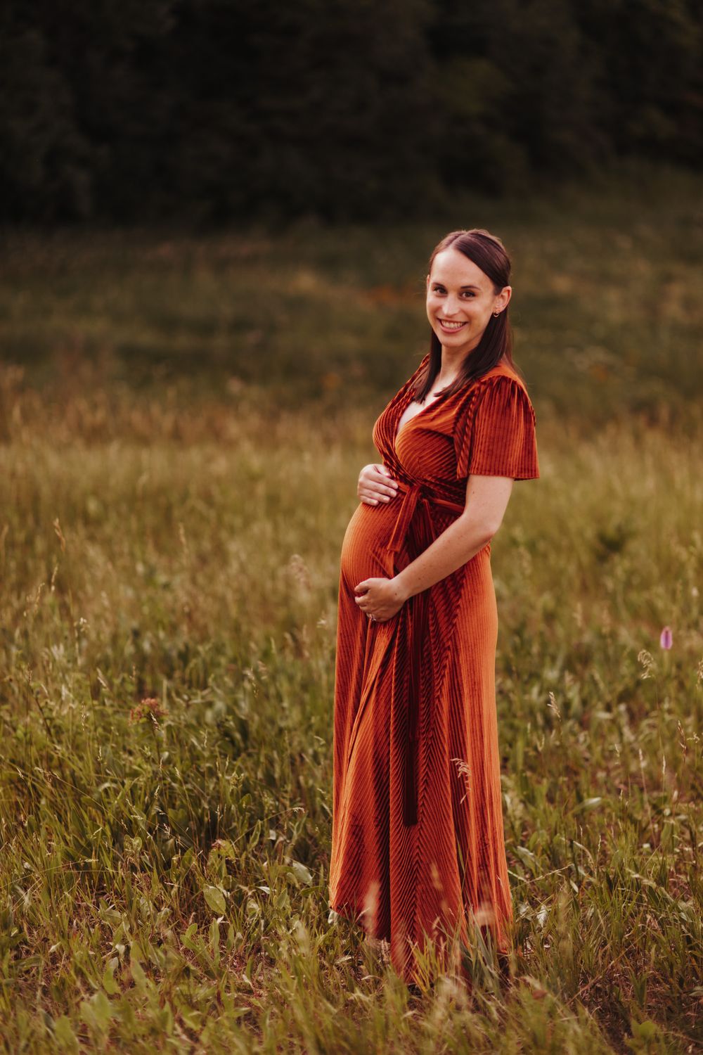 Pregnant woman in a burnt orange dress smiles at the camera as she stands in a field of green tallgrass.