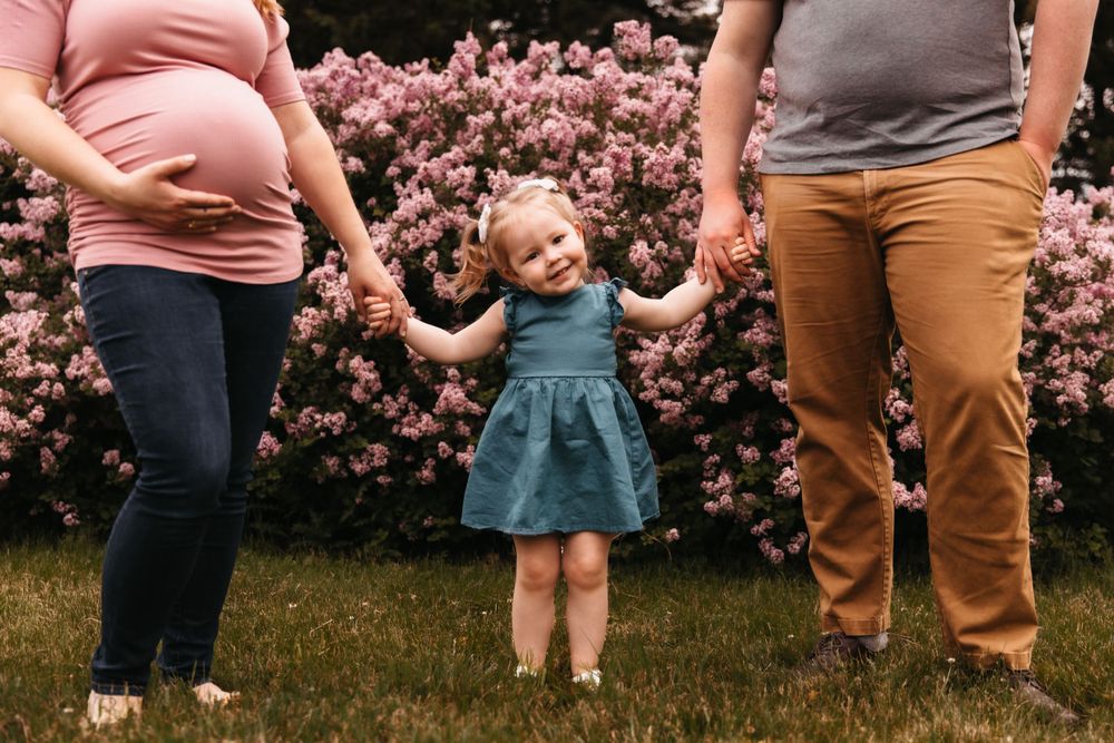 Pregnant woman and her husband hold their toddler's hands. The toddler smiles at the camera.