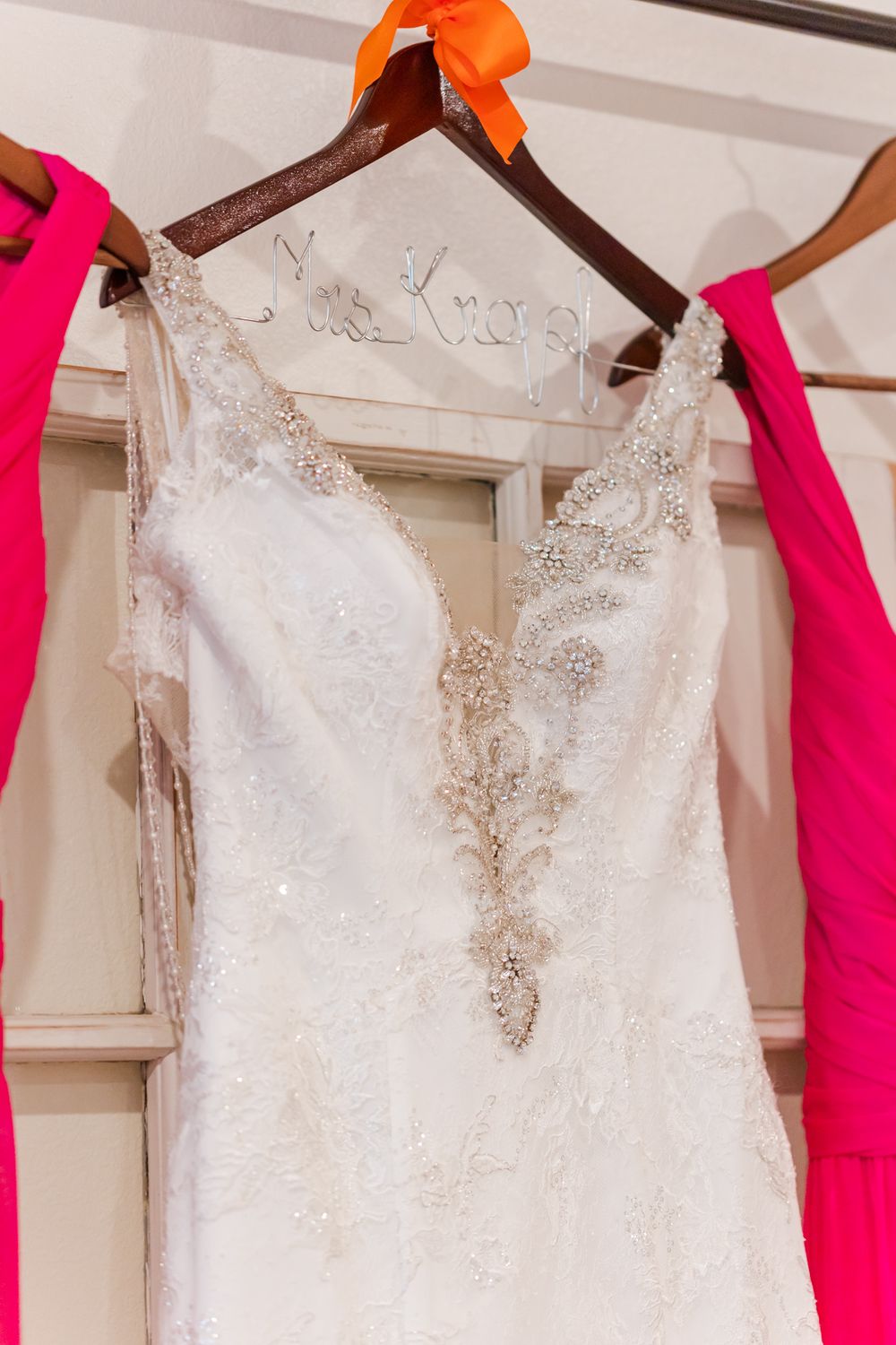 White lace wedding dress with intricate beading displayed on wooden hanger with orange ribbon.
