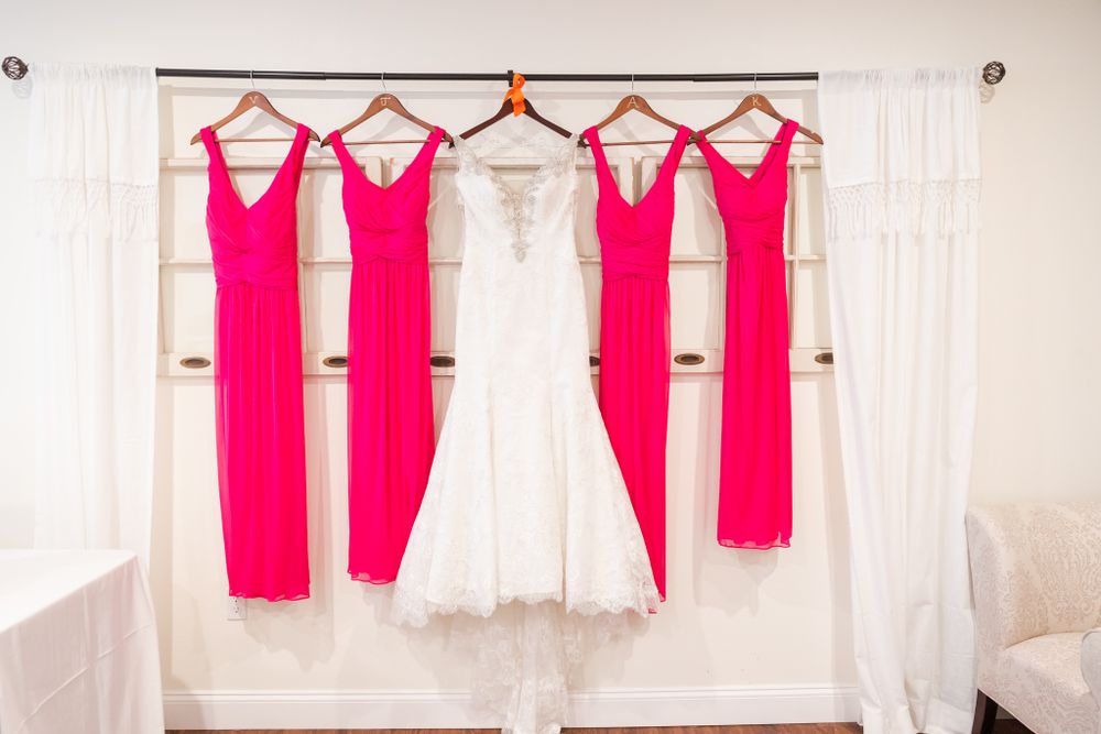 Pink bridesmaid dresses and a white wedding gown hanging on a rack against a light backdrop.