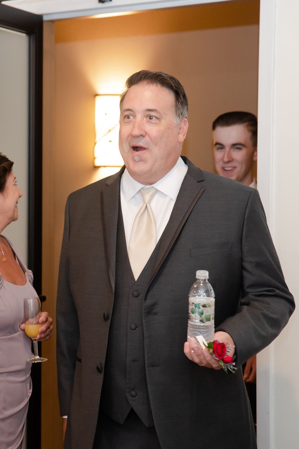 Wedding party member in formal attire holding a water bottle and boutonniere.