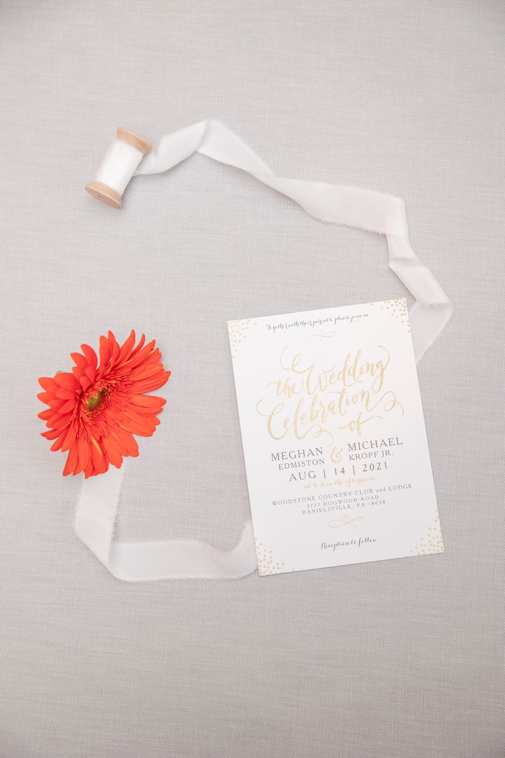 Wedding invitation with gold lettering displayed with coral gerbera daisy and white ribbon.