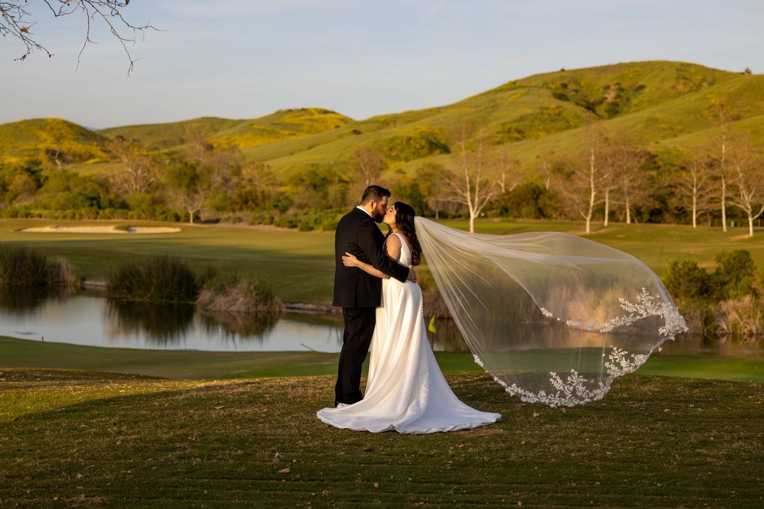 Newlyweds embracing on a grassy hill by a lake with a flowing veil at sunset.