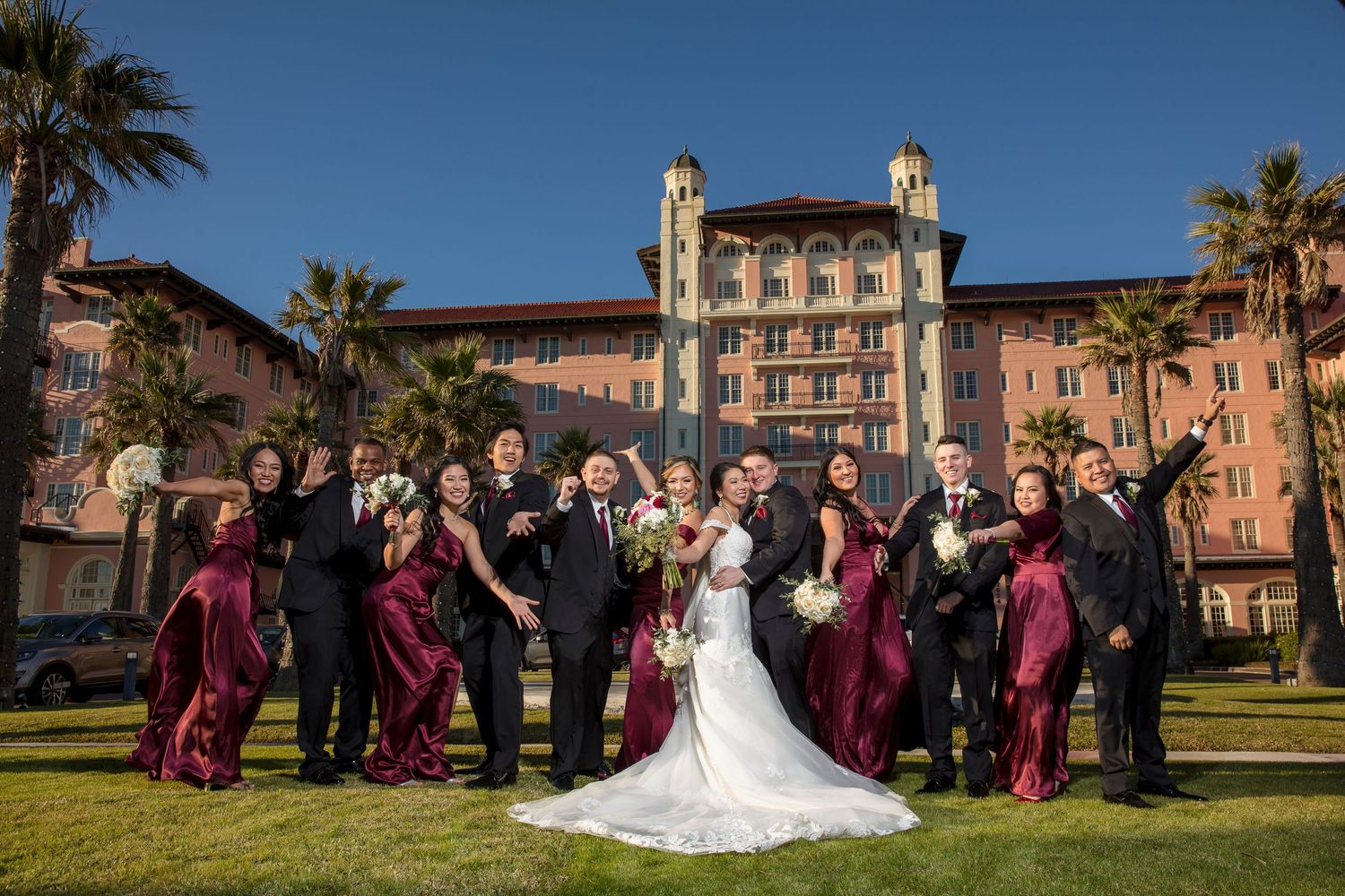 Wedding party gathered outside historic pink Mediterranean-style building with palm trees.