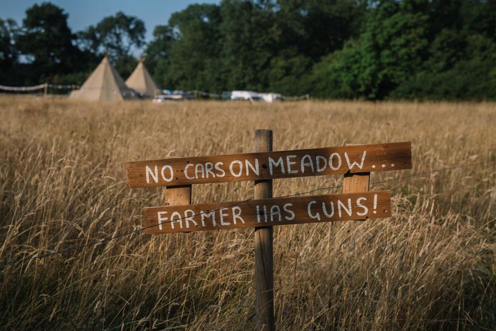 Wooden sign in meadow reads 'No Cars on Meadow, Farmer Has Guns!' with tipis in background.