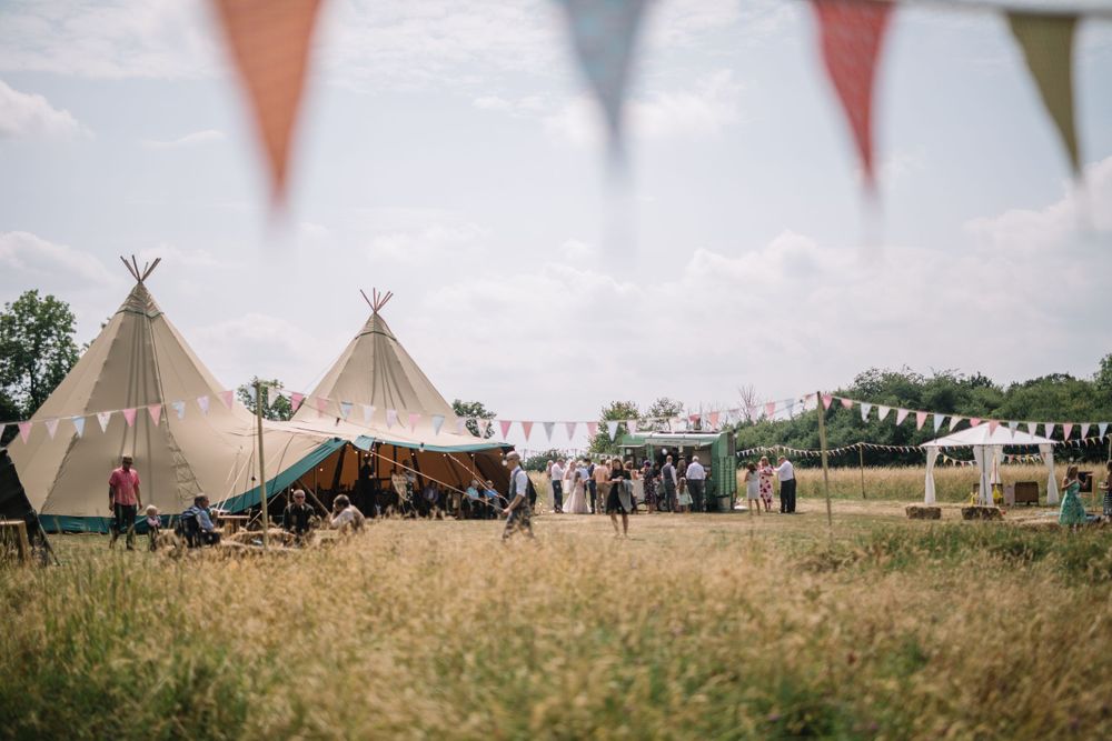 Rustic outdoor wedding venue with teepee tents and decorative bunting flags in a grassy field.