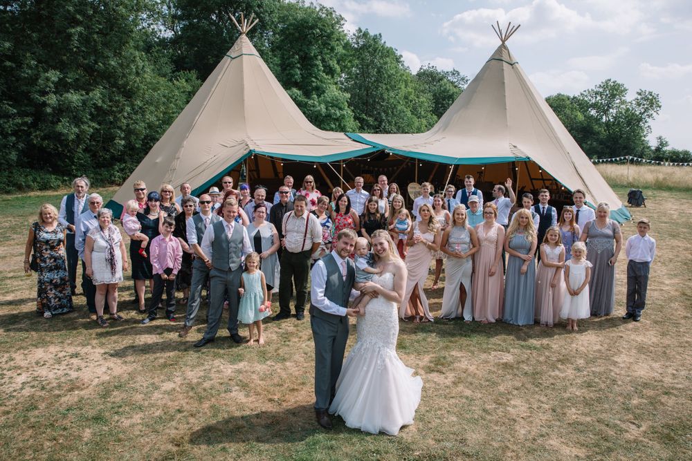 Large wedding group gathers outside teepee tent reception venue on sunny day.