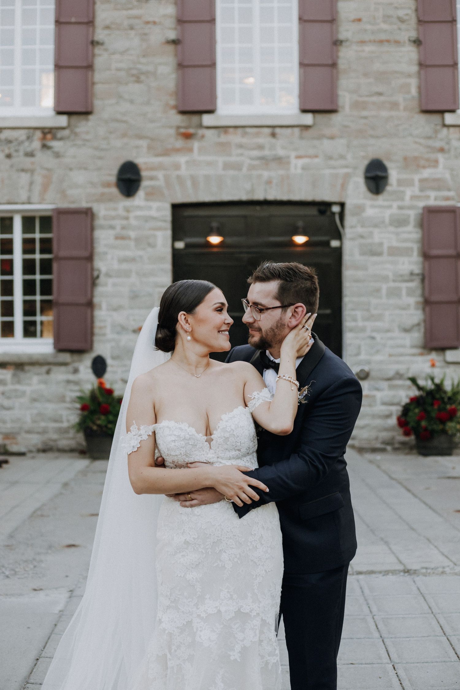 Wedding couple sharing intimate moment outside stone building with burgundy shutters.